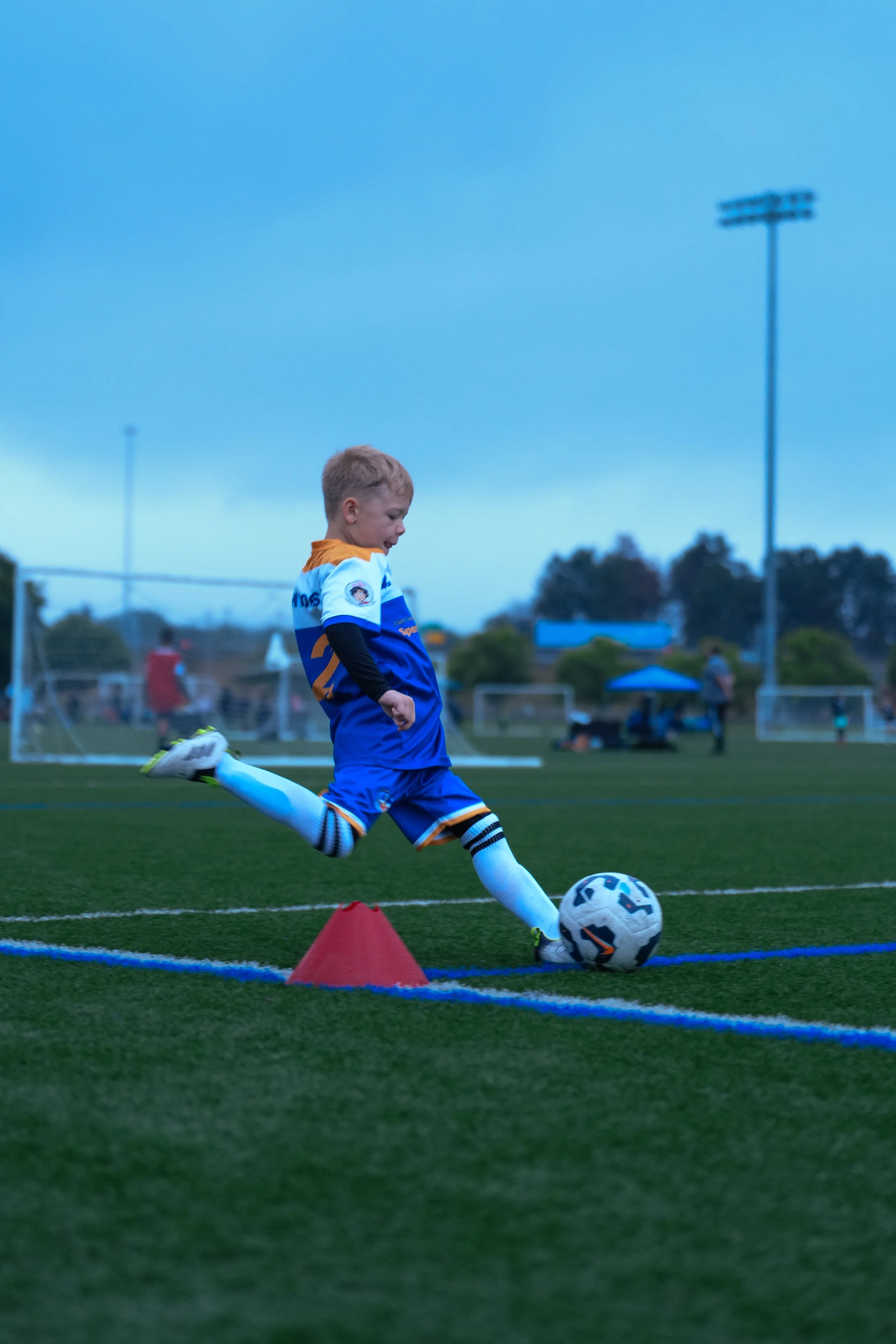 Young boy in blue and orange soccer uniform kicking a soccer ball during practice on a grassy field, with cones set up and cloudy sky overhead.