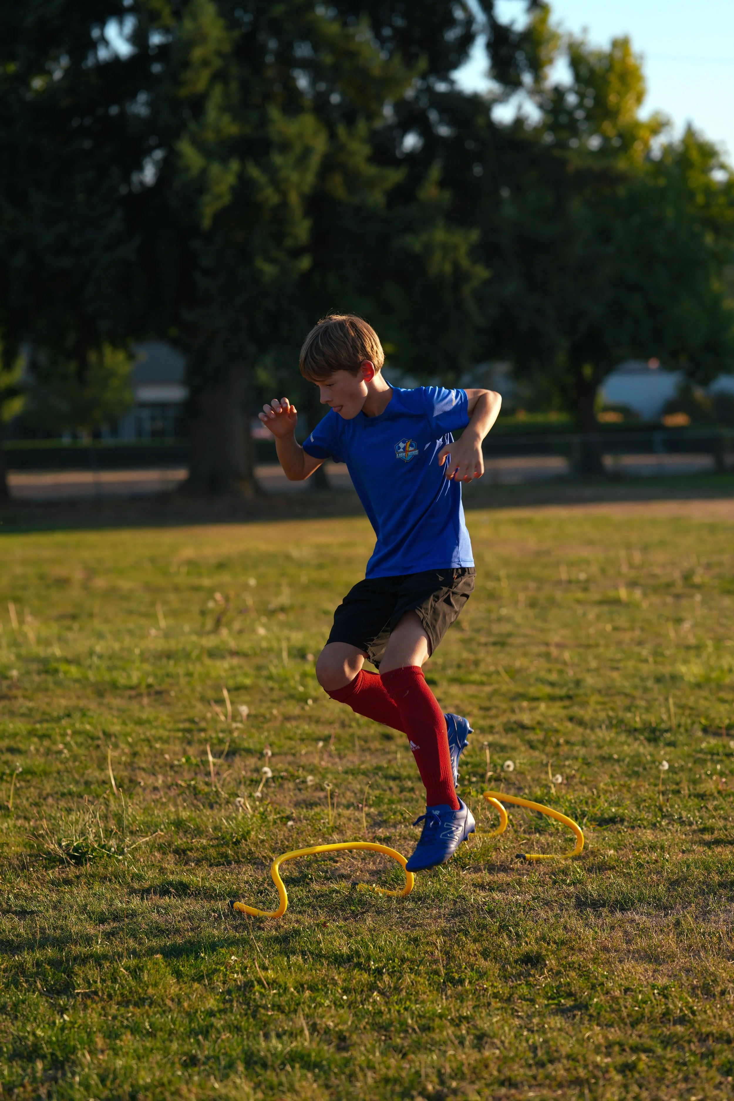 Young boy wearing a blue shirt, black shorts, red socks, and blue cleats jumping over a yellow agility hurdle on a grassy field in the late afternoon.