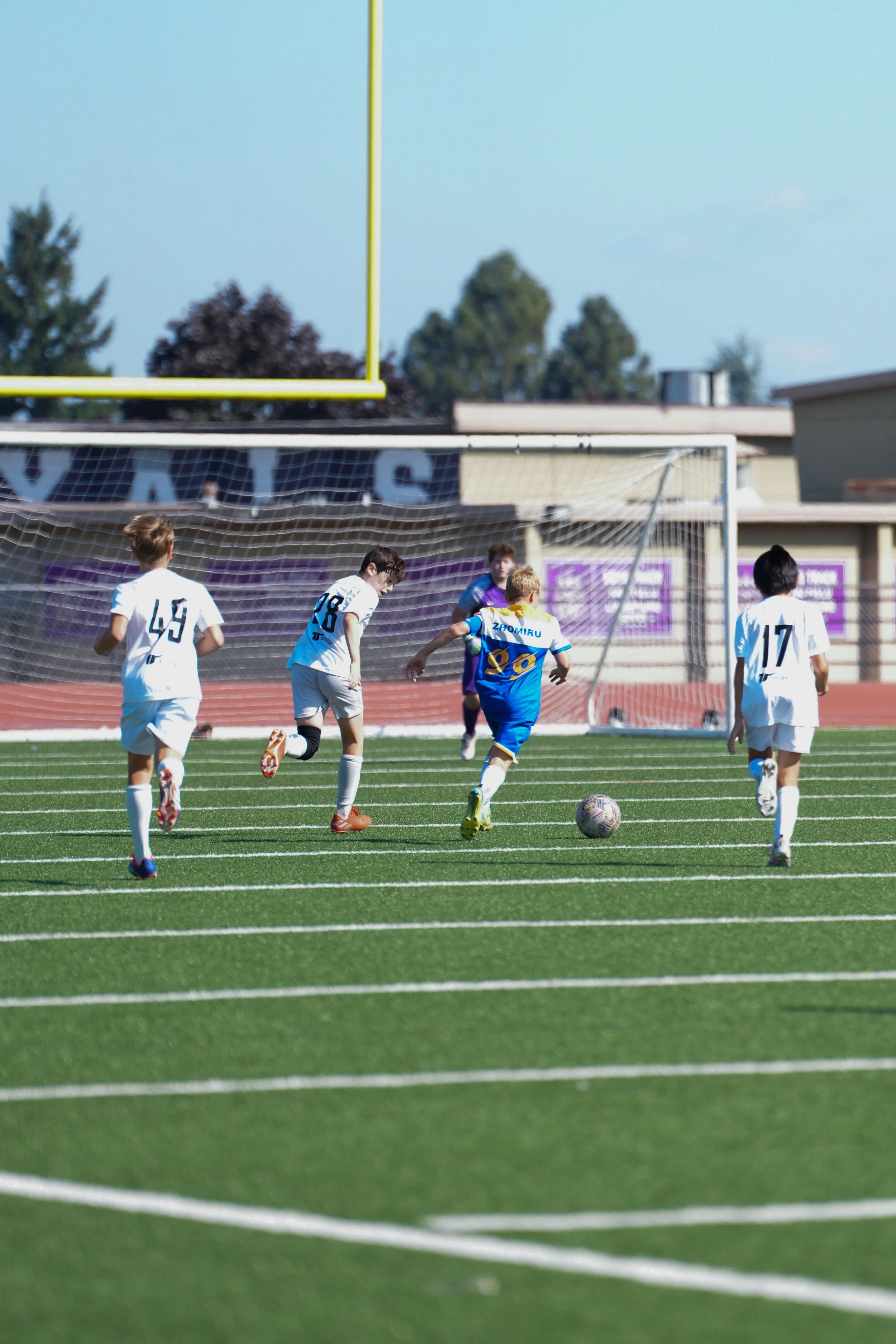 Young kids playing soccer on a green field with a goal and net in the background, during a sunny day.