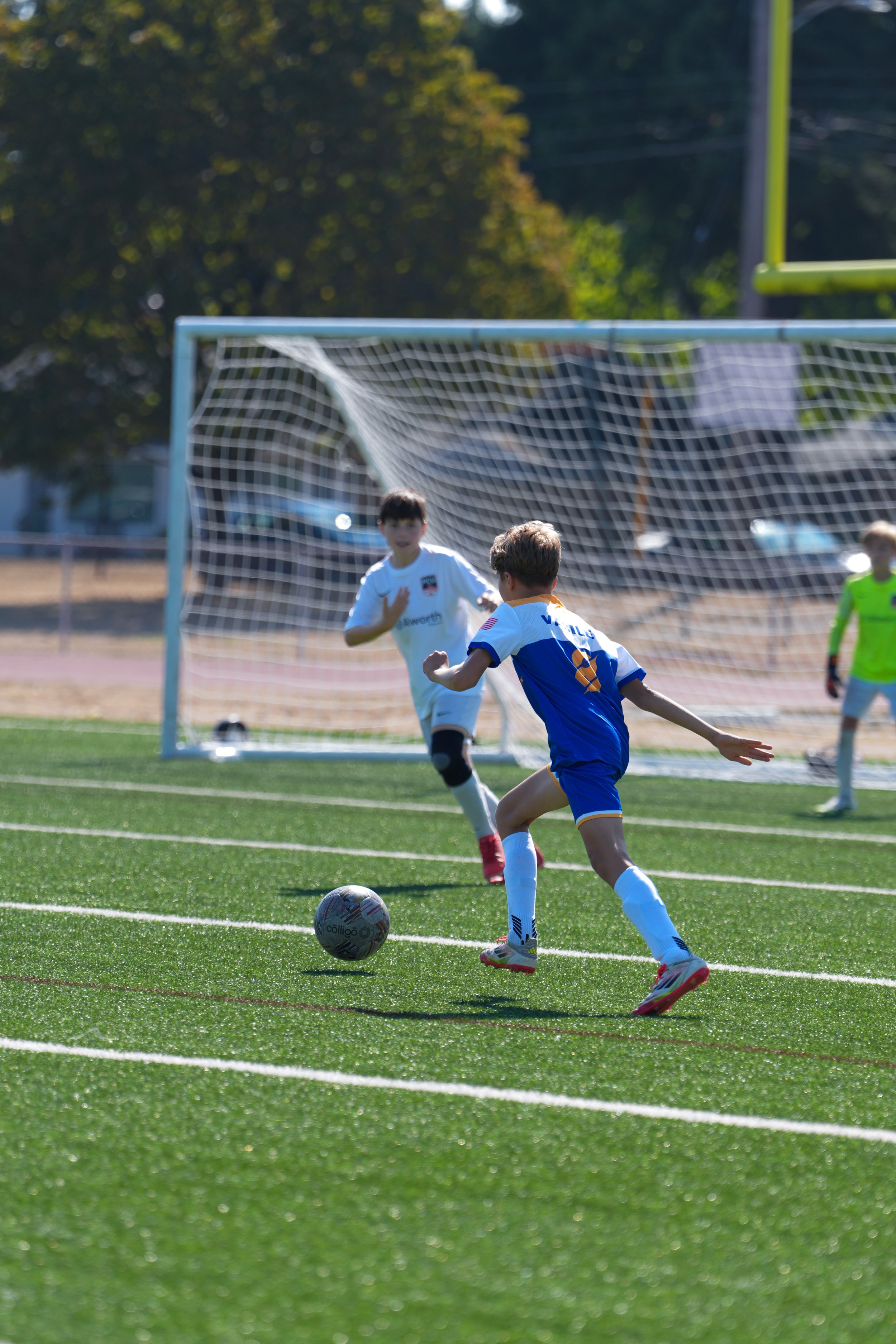 Children playing soccer on a field, with one child about to kick the ball towards the goal while another child in a white jersey is near the goal. The goalkeeper is in the background.