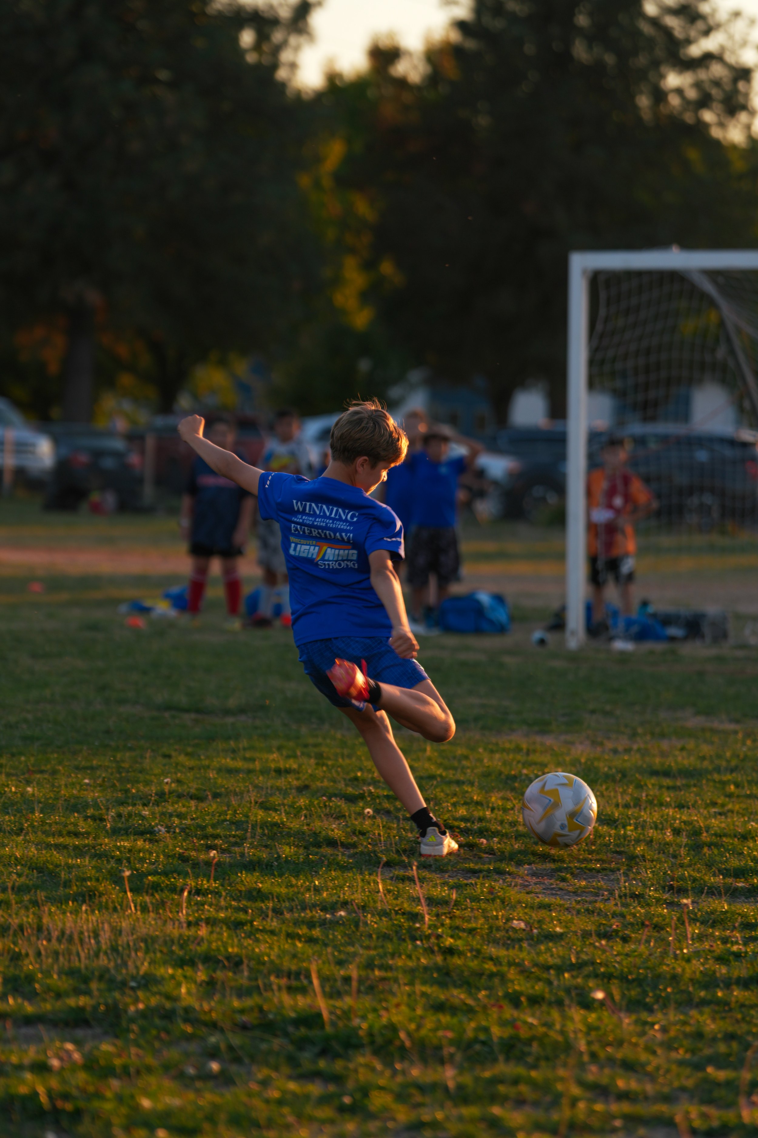 A young boy in a blue T-shirt and shorts kicking a soccer ball on a grassy field during sunset, with a goalpost and trees in the background.