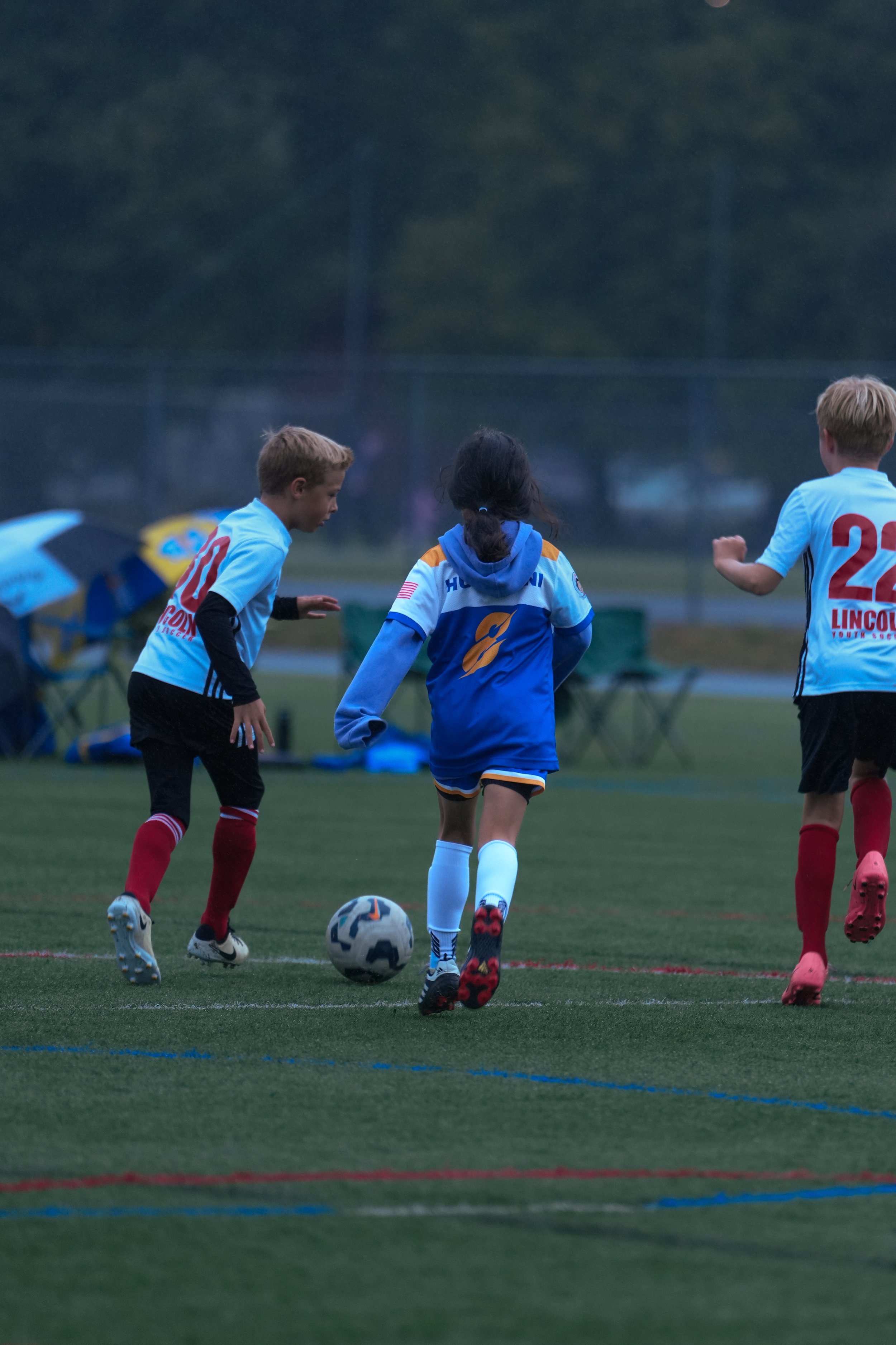Kids playing soccer on a field, two wearing light blue jerseys and one in a darker blue hoodie, with a soccer ball in the foreground.