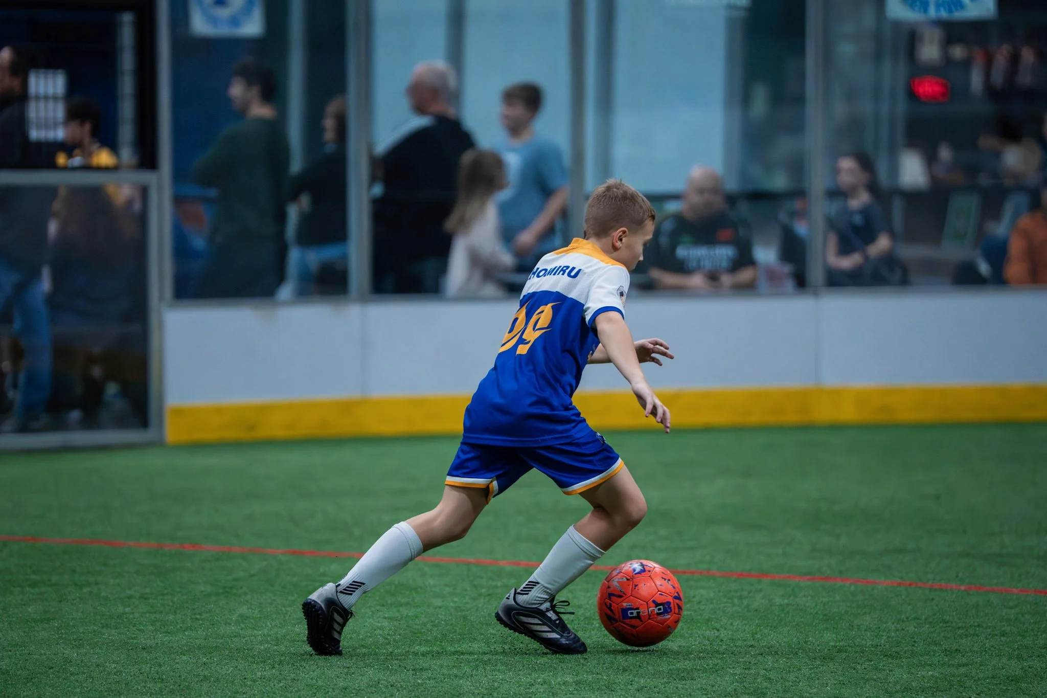 A young boy in a blue and white soccer uniform dribbling a red soccer ball on an indoor field, with spectators in the background watching behind glass.