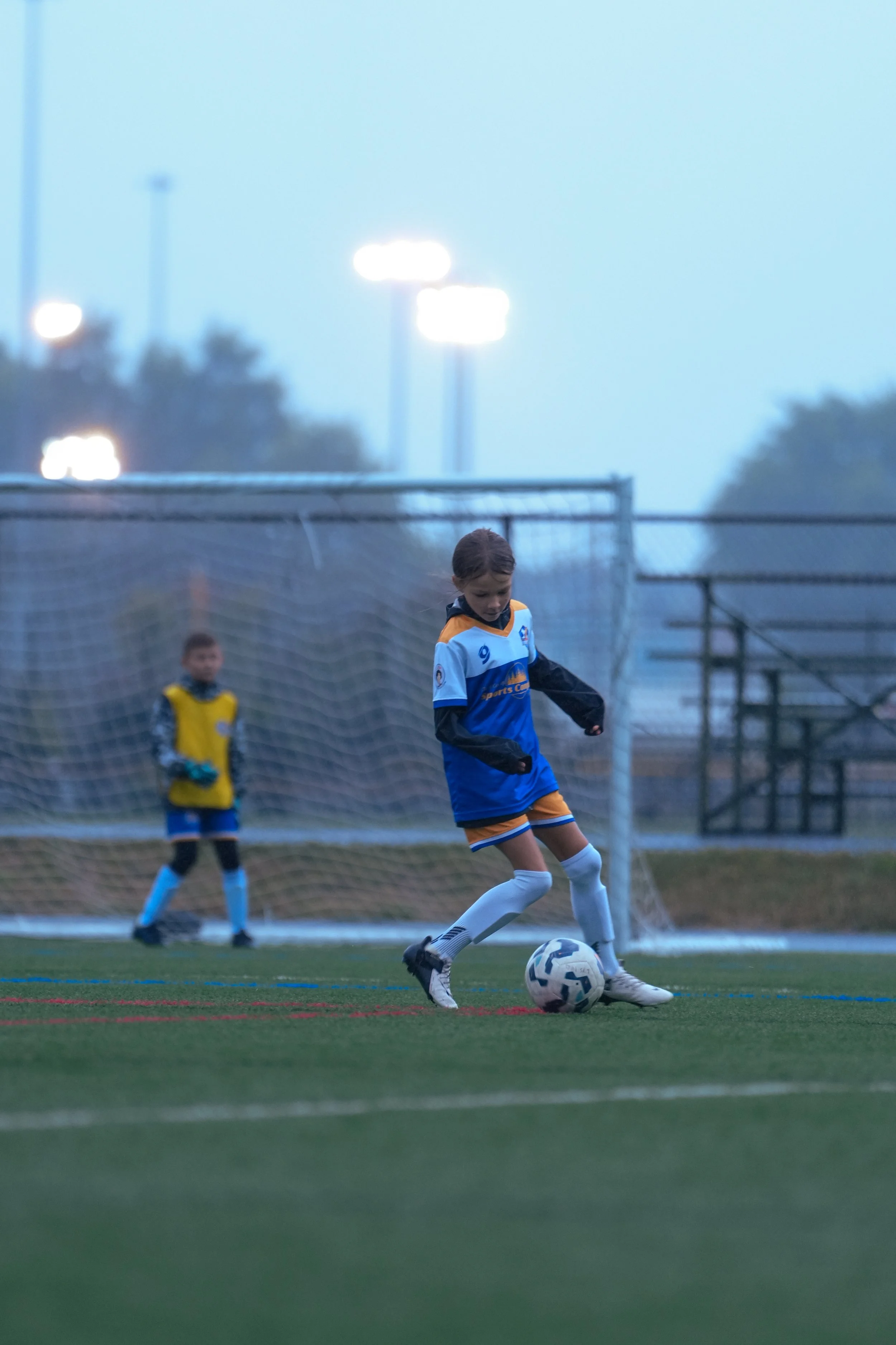 Young girl in a blue and orange soccer uniform kicking a soccer ball on a field, with a goalkeeper in a yellow jersey and a goalpost in the background during dusk.