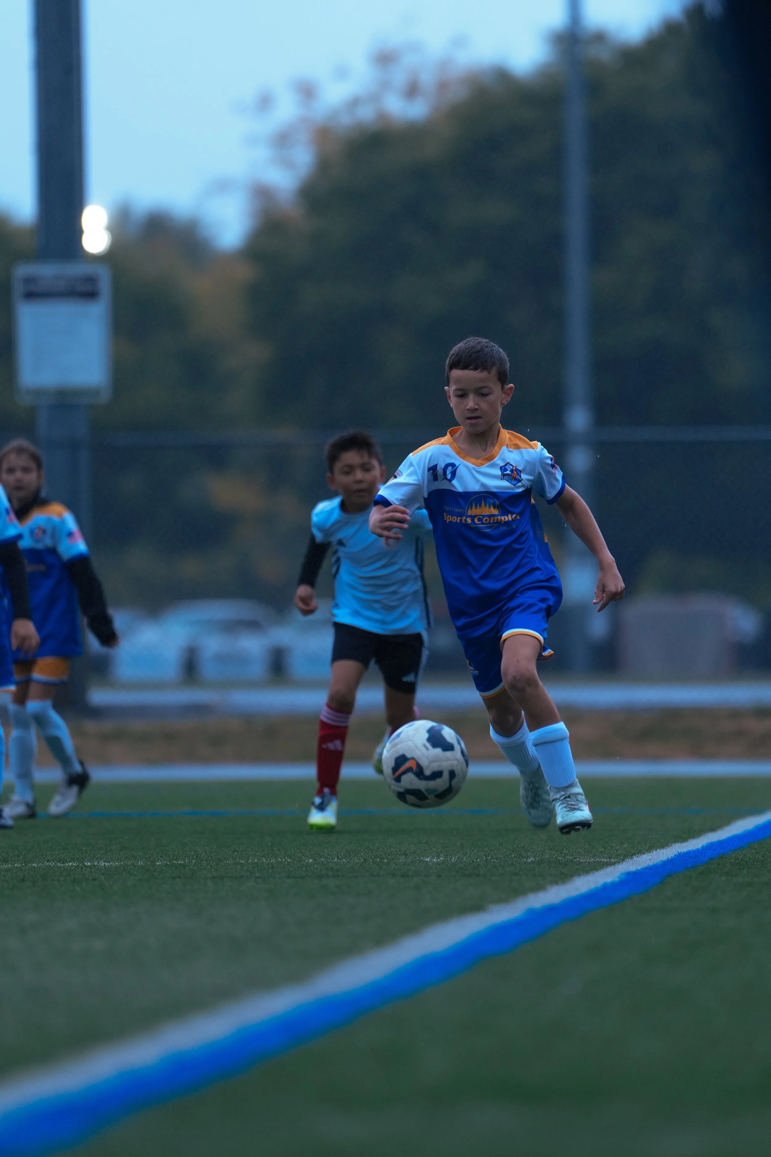 Children playing soccer on a field during dusk, with one boy kicking the ball while others run behind him.
