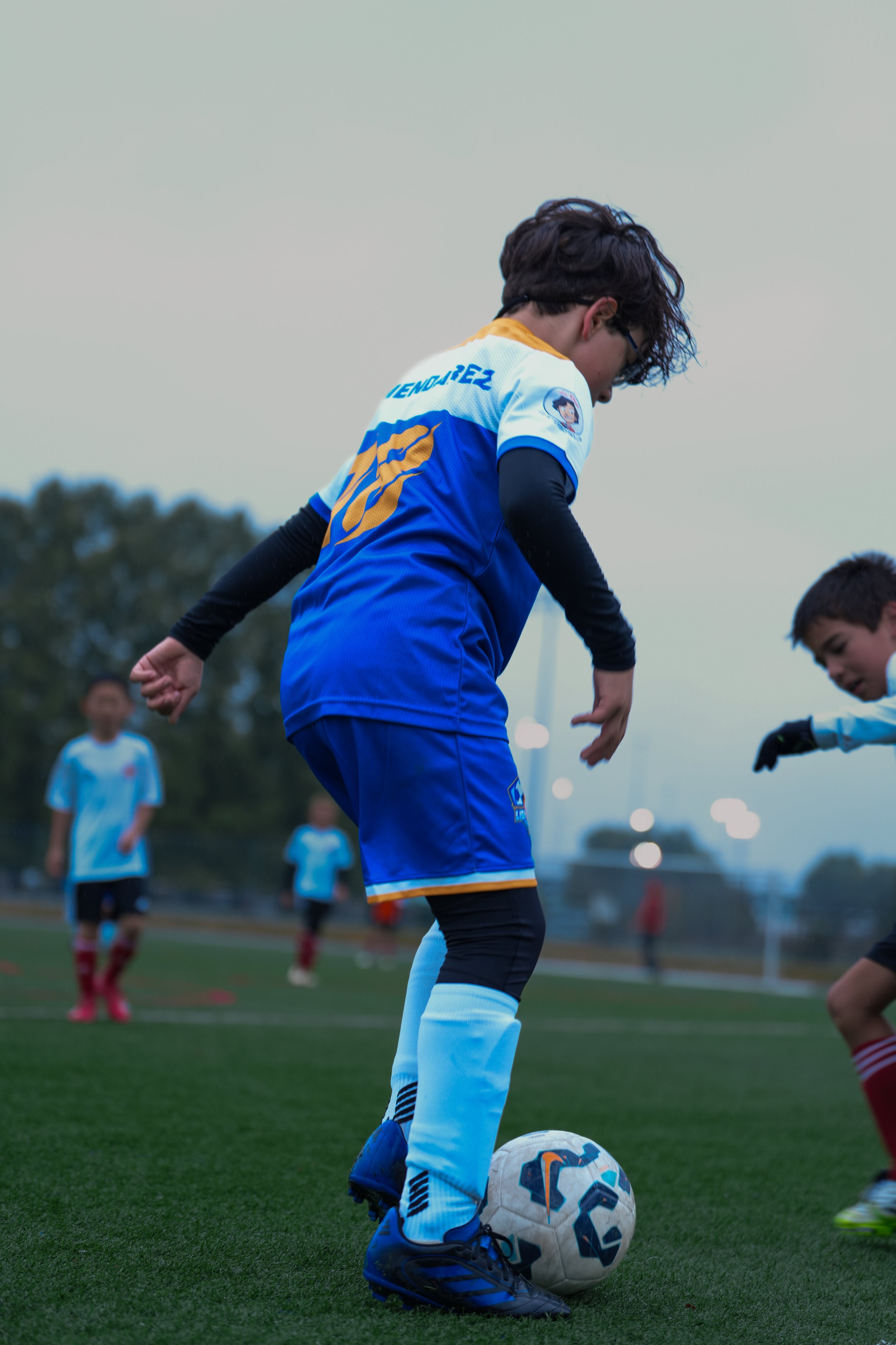 Young soccer player with glasses and wearing a blue and white uniform, standing on a soccer field with a ball, surrounded by other children playing soccer.