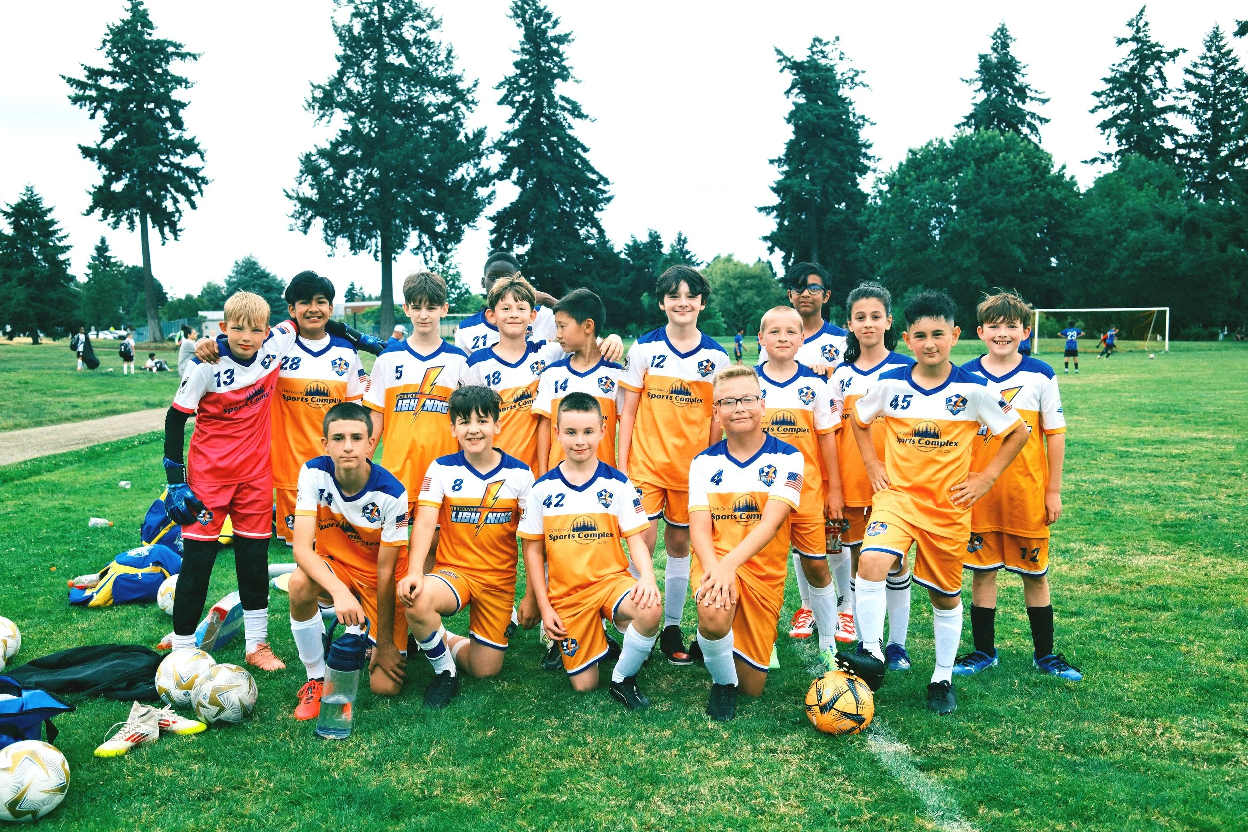 A group of young boys and girls in soccer uniforms standing on a soccer field, posing for a team photo with trees and a goal post in the background.