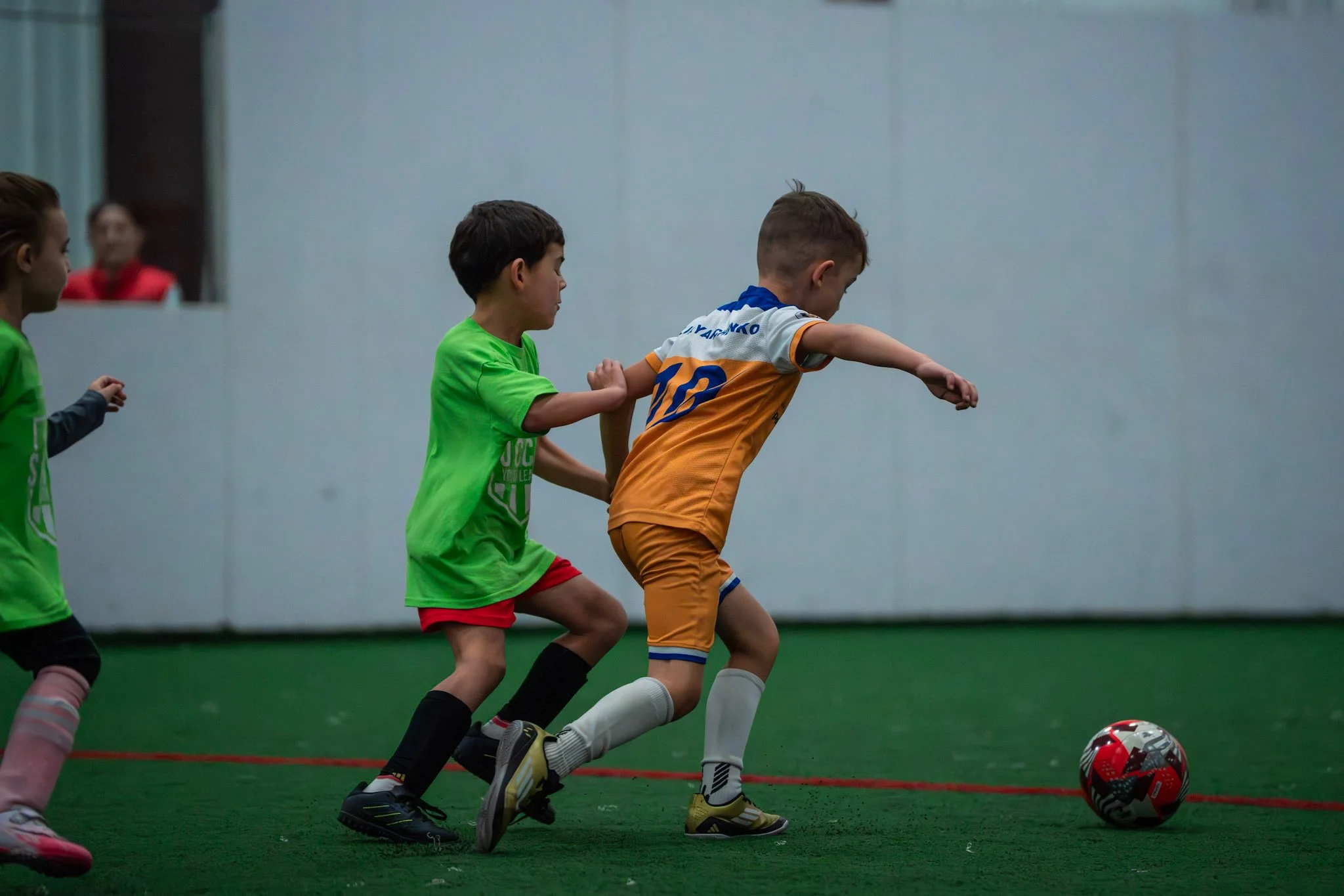 Children playing indoor soccer, chasing the ball on green artificial turf.