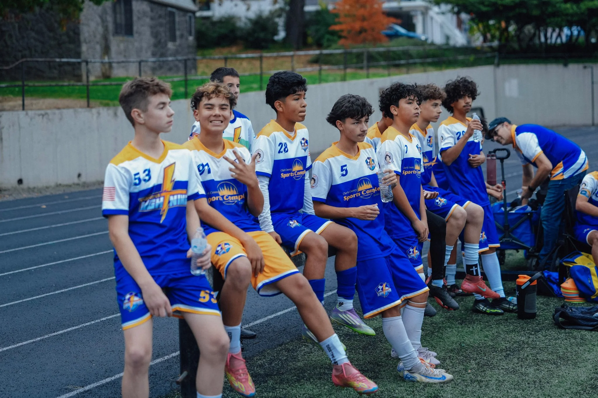 Young soccer players sitting on a bench during a game, dressed in white and blue jerseys, holding water bottles.
