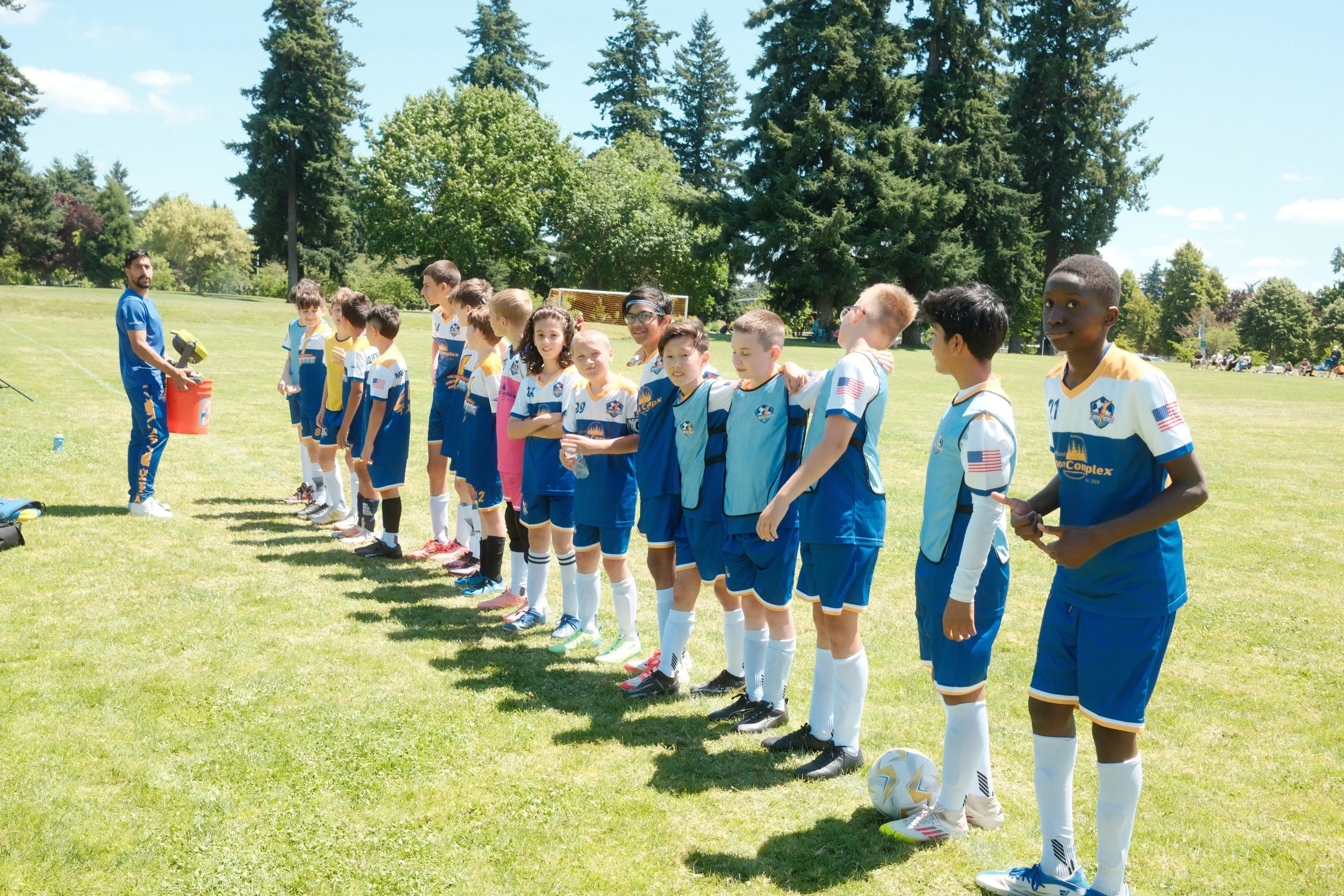 Children in soccer uniforms lining up on a grassy field as a coach gives instructions, with trees and a blue sky in the background.