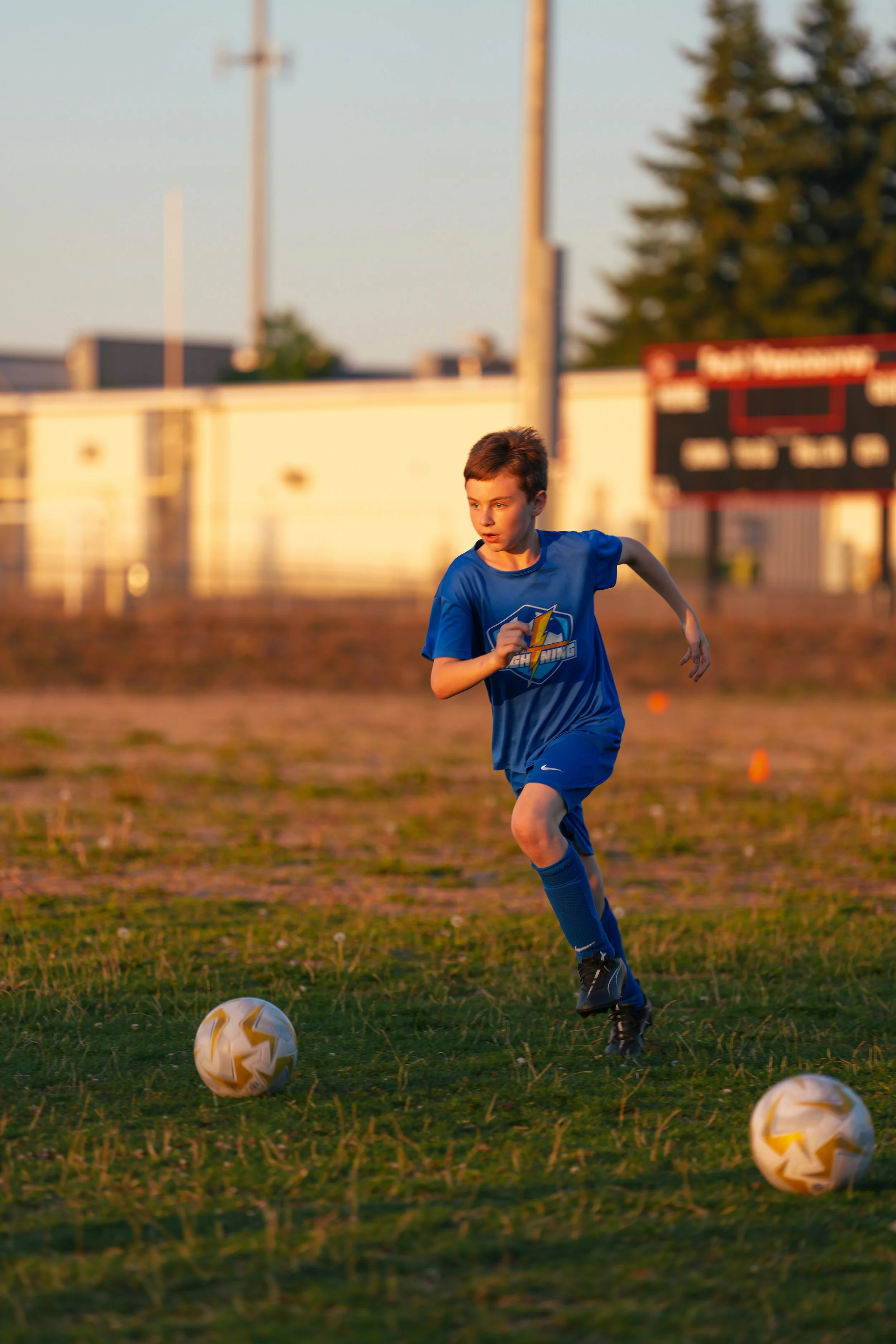 A young boy in a blue soccer uniform is running on a soccer field during sunset, with two soccer balls in front of him and a blurred scoreboard and trees in the background.