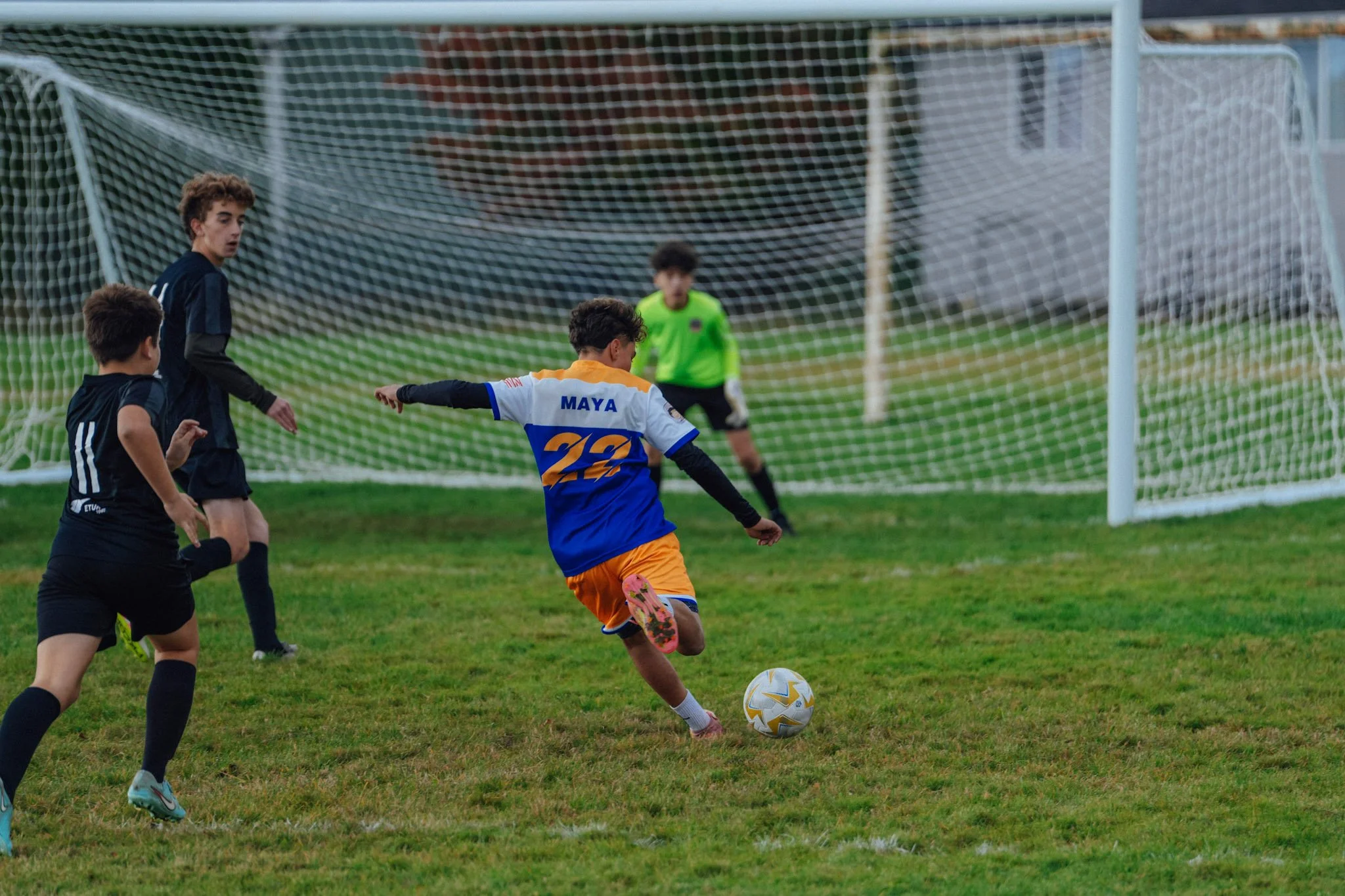Young soccer players on a grass field, one in orange and blue kicking the ball towards the goal, with a goalkeeper in green in the background, and two other players watching.