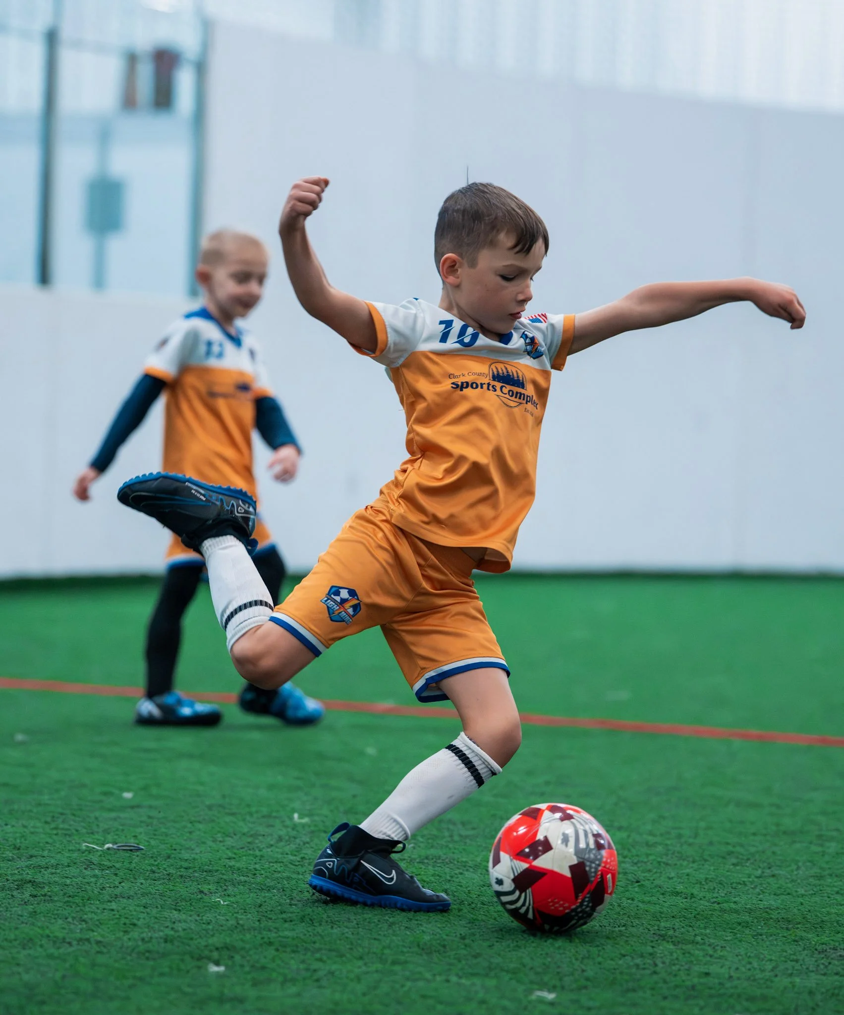 Two young boys playing indoor soccer; one boy kicking a ball while running, wearing an orange uniform, and a girl in the background wearing a similar uniform.