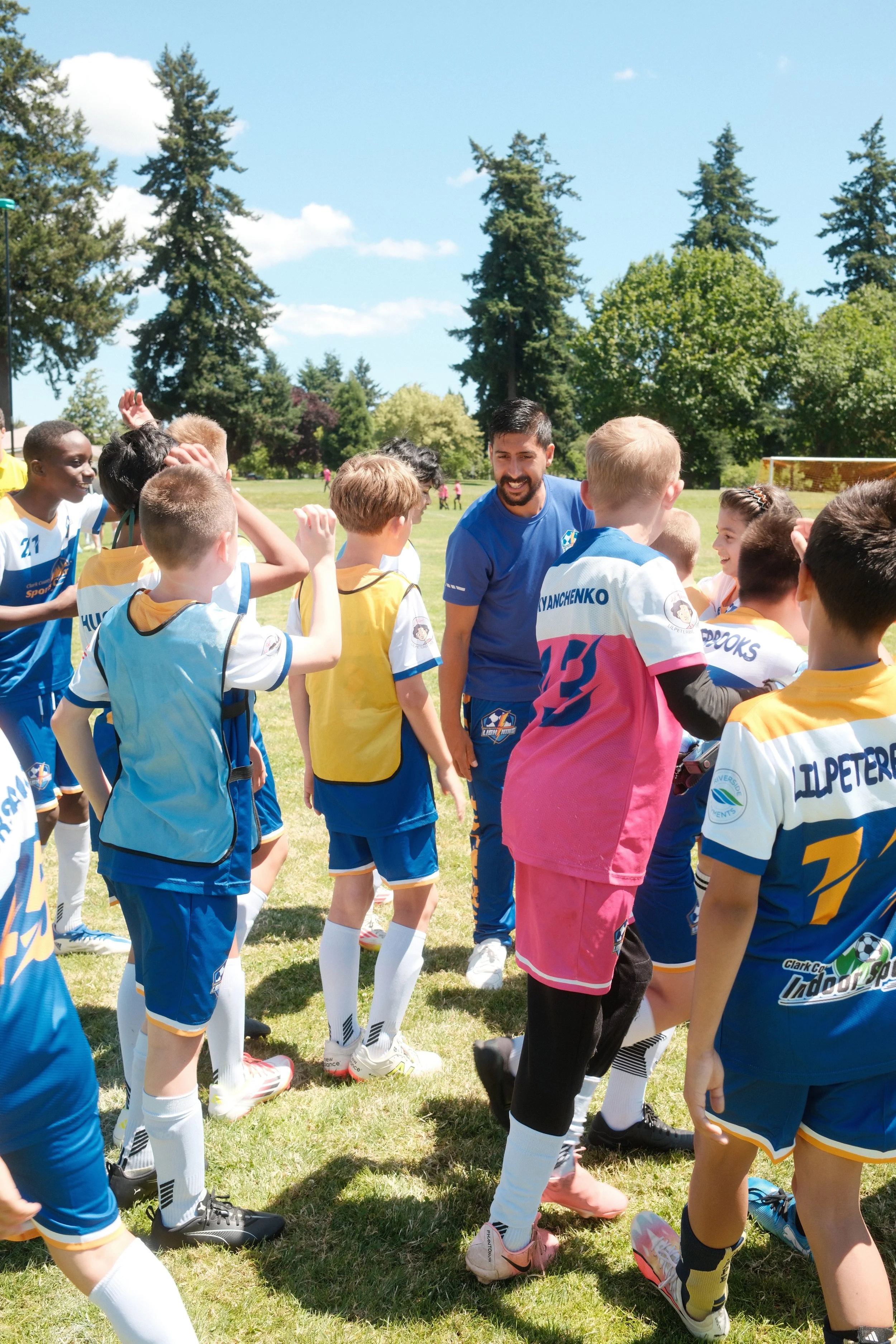 Children in soccer uniforms gather around a coach during a game on a field under a blue sky with trees in the background.