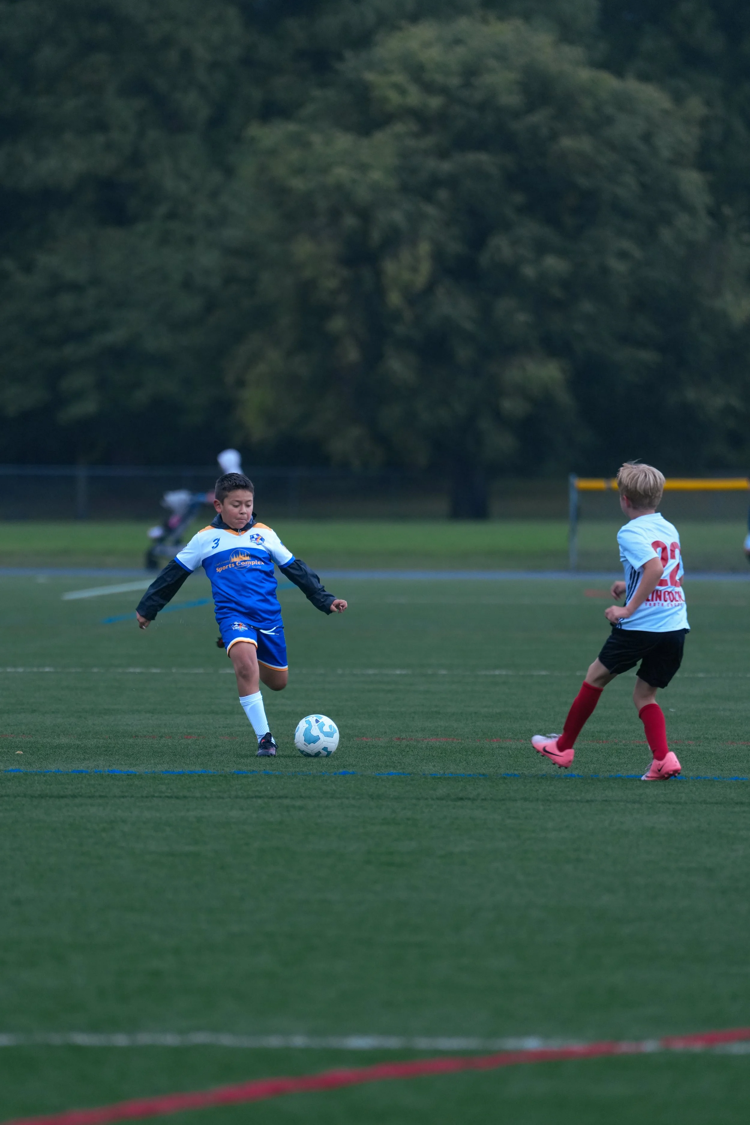 Two young boys playing soccer on a field, with one boy about to kick the ball and another boy watching.