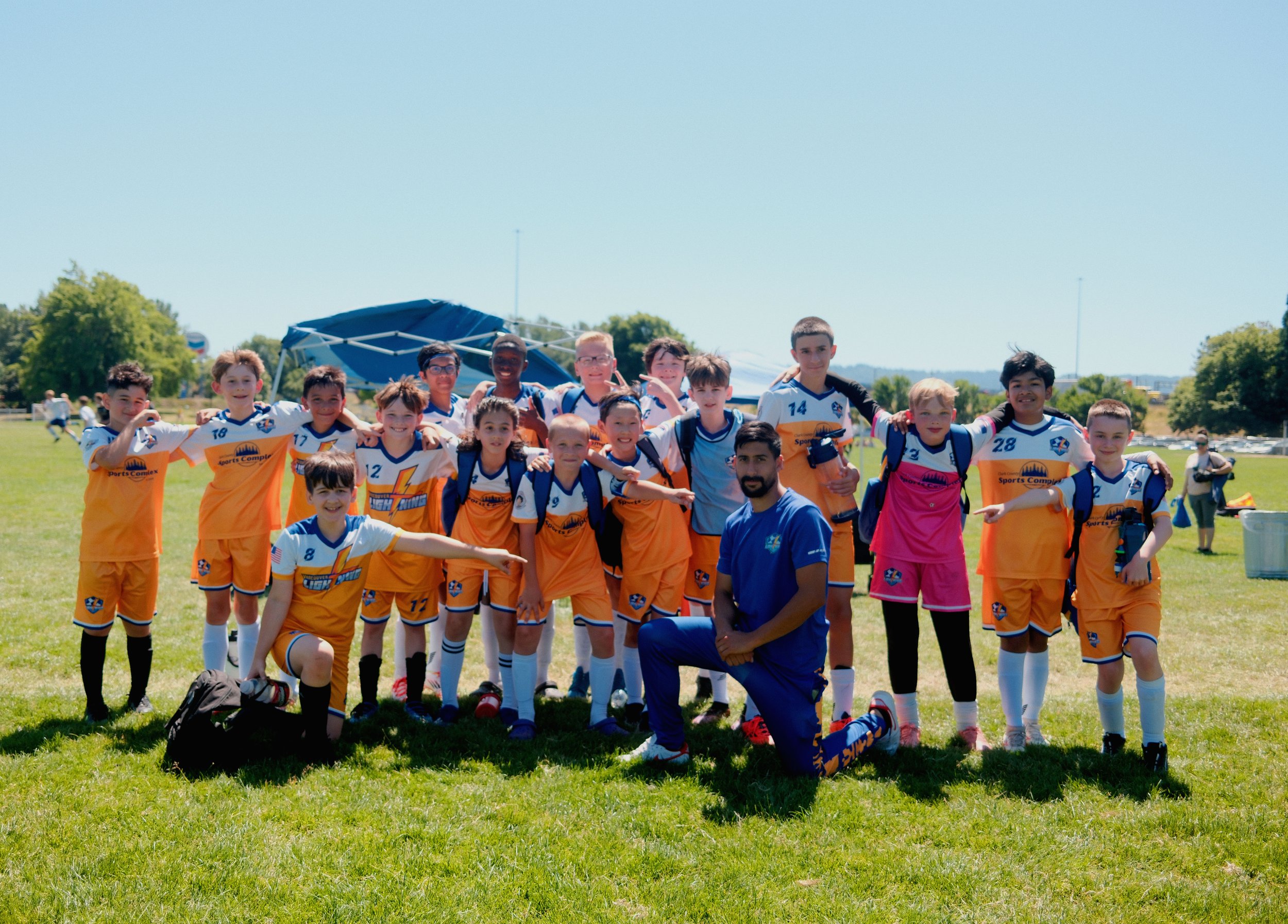 A youth soccer team of children and their coach pose on a grassy field after a game or practice. The team is wearing matching orange and white uniforms with blue accents, and some children are wearing backpacks. The coach is kneeling in front and to 