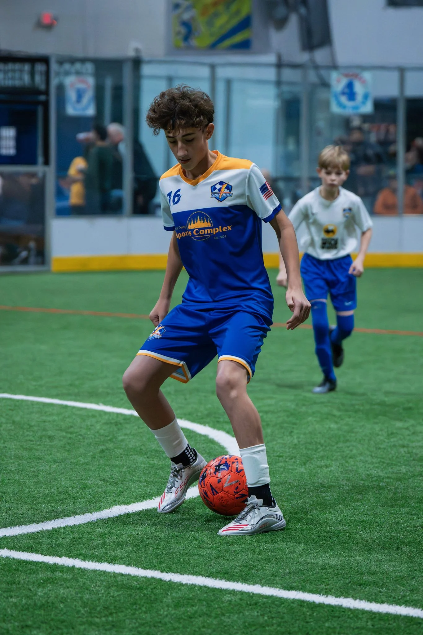 Young boy in blue and white soccer uniform playing indoor soccer on green turf, with another child in the background.