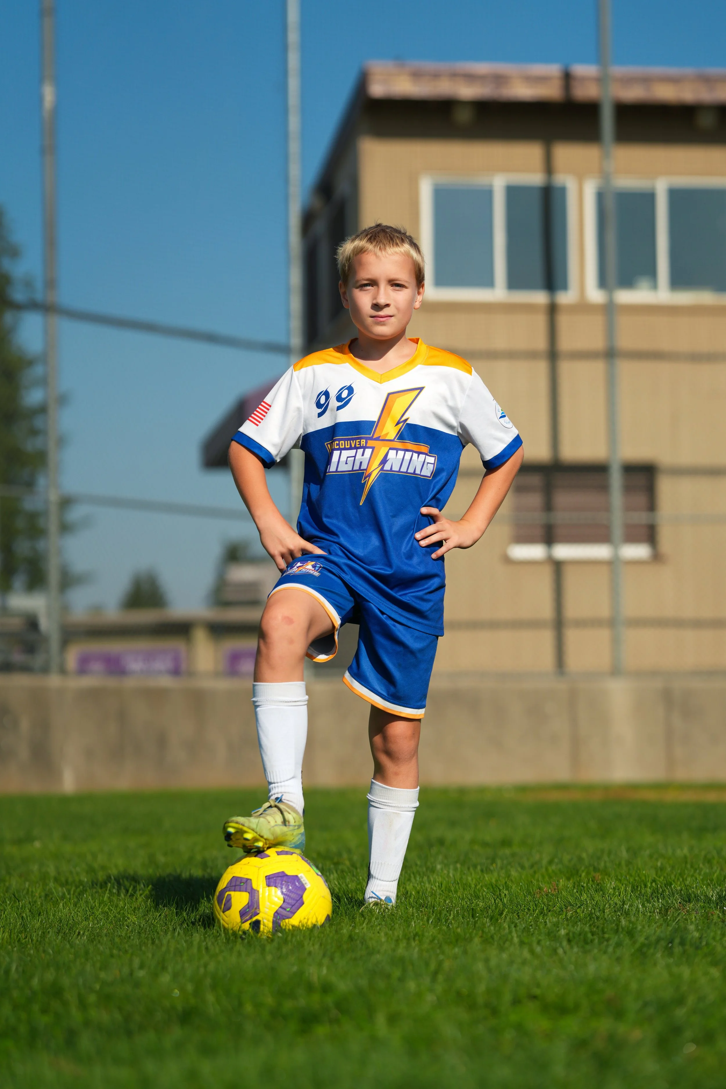 A young boy in a soccer uniform standing on a soccer field, with one foot on a yellow and purple soccer ball, posing confidently with his hands on his hips.