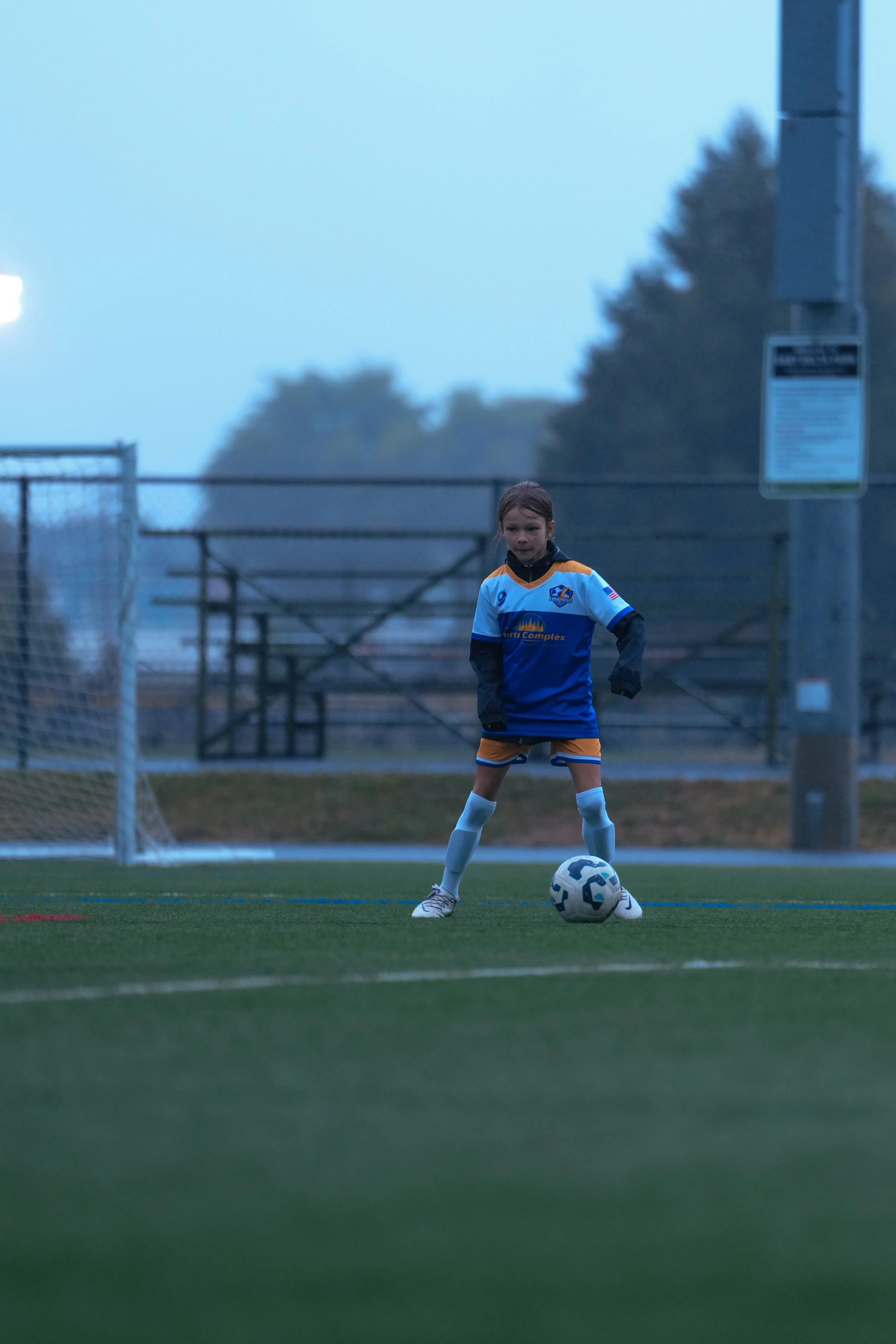 A young girl wearing a sports uniform stands on a soccer field with a ball at her feet, preparing to kick.