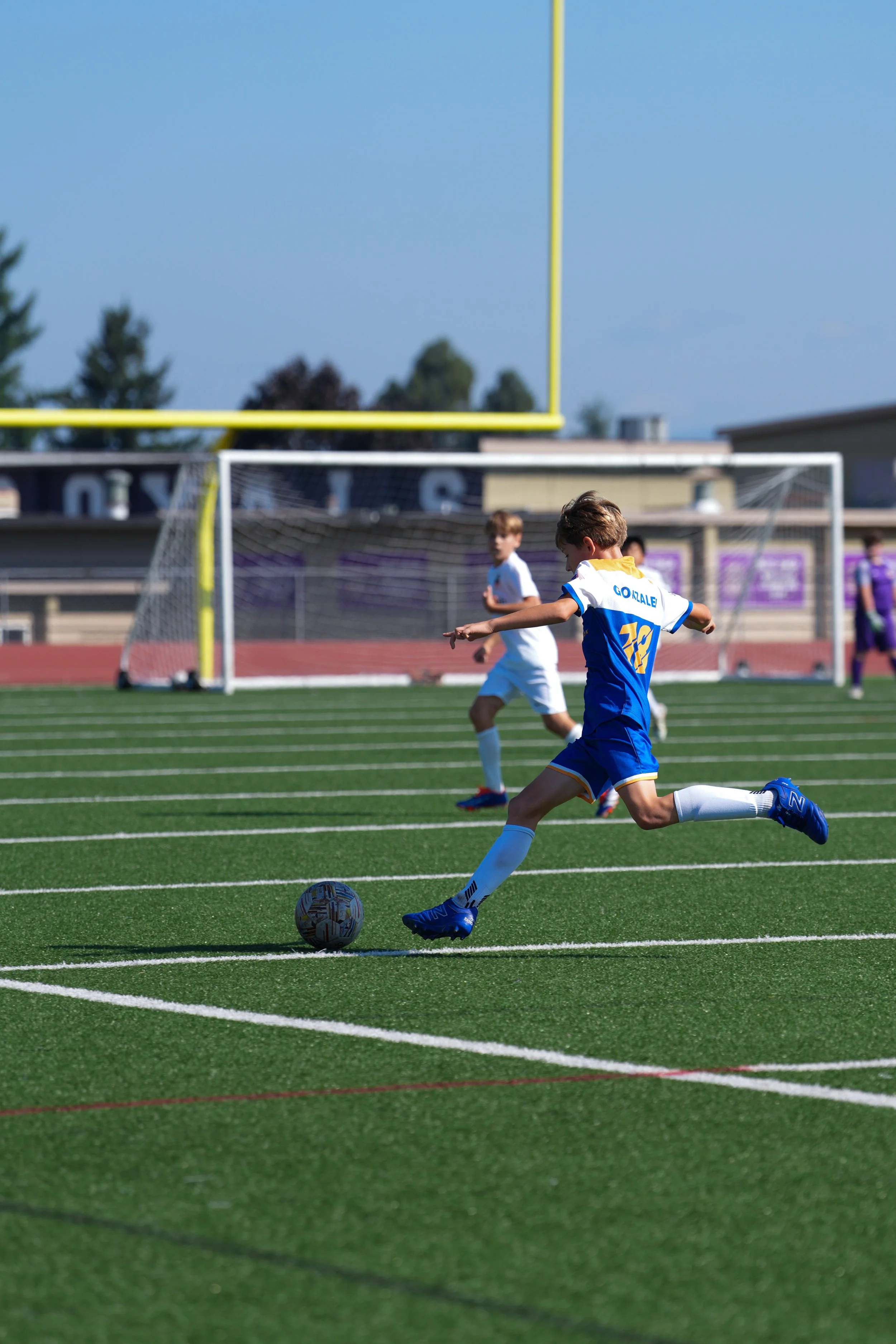 Young soccer player in blue and white uniform kicking a soccer ball on a field with goal in background.