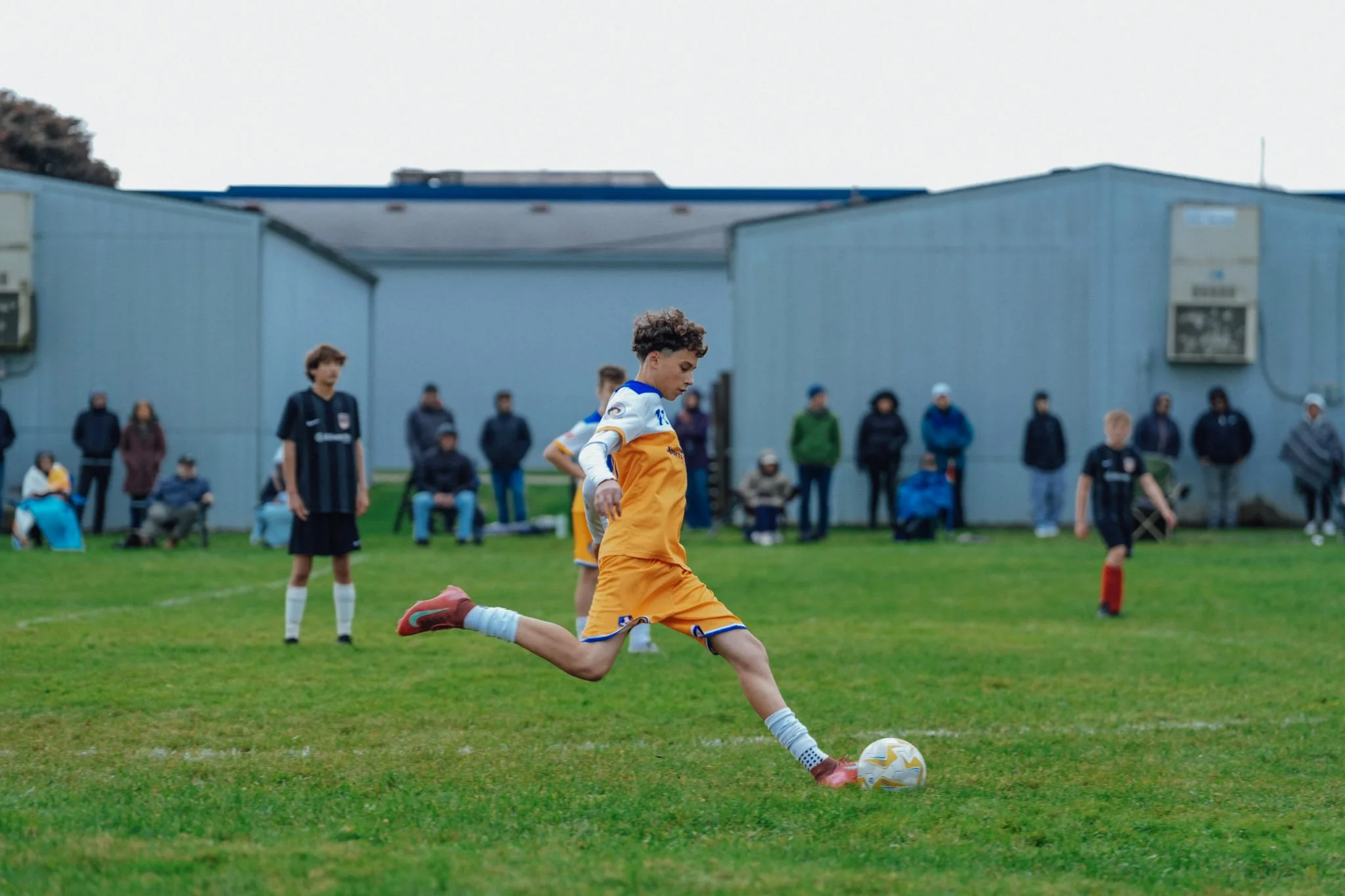 A young soccer player in a yellow and white uniform kicks a ball on a grass field, with spectators watching from the sidelines.