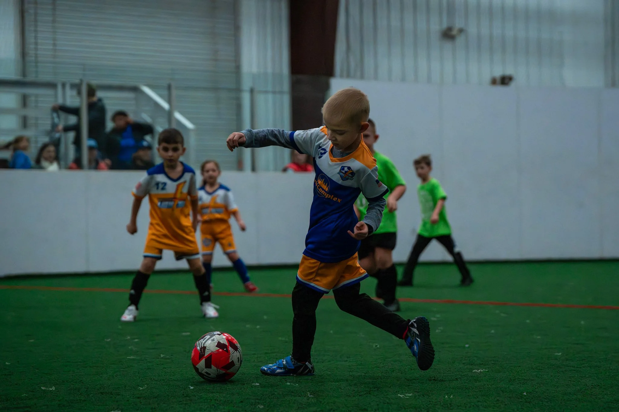 Children playing indoor soccer on a green field, with one boy kicking a soccer ball while others watch.