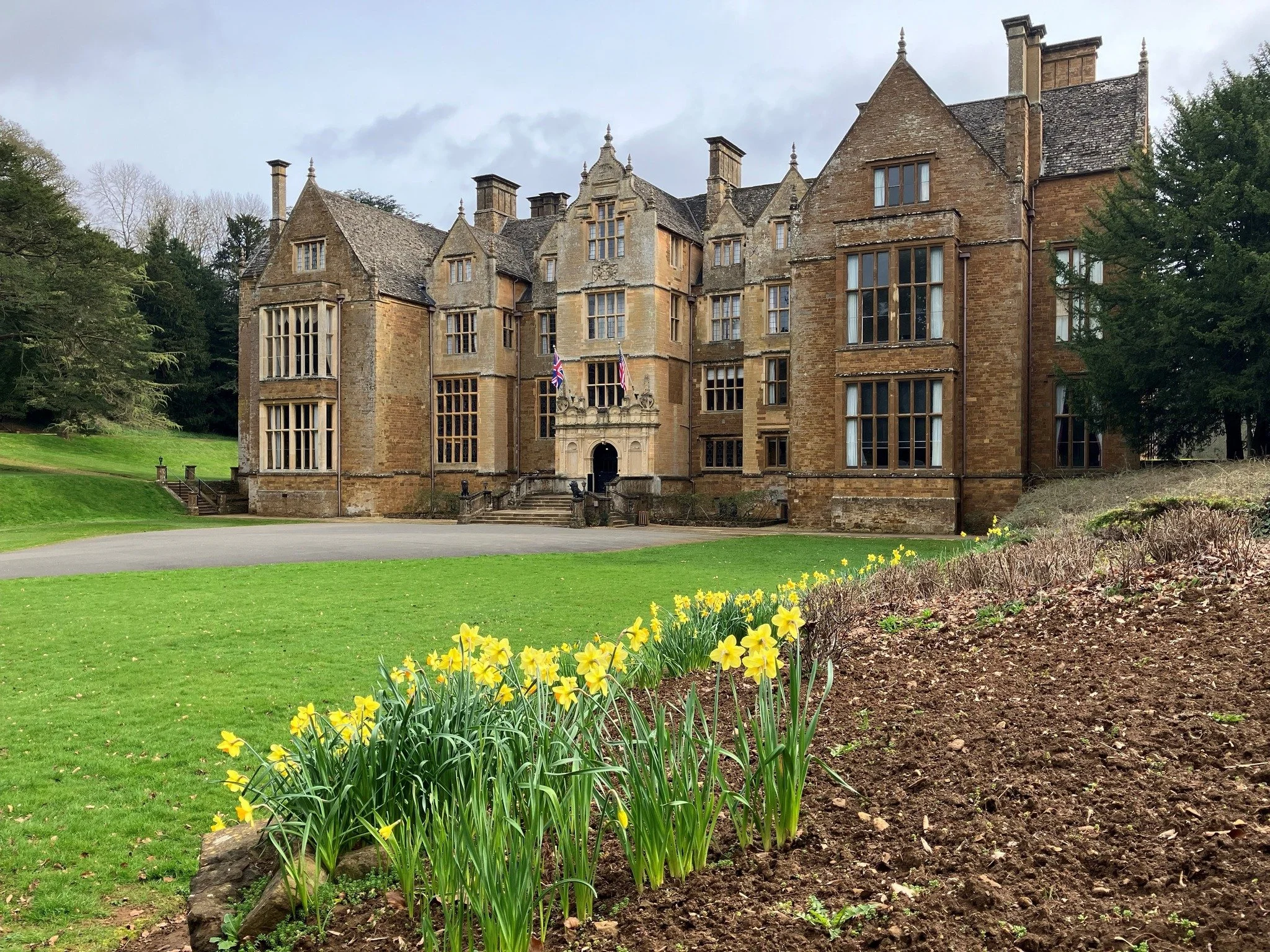 Wroxton Abbey, a large, historic stone house or castle with multiple windows, flags near the entrance, surrounded by green lawns, trees, and flowerbeds with yellow flowers in the foreground.
