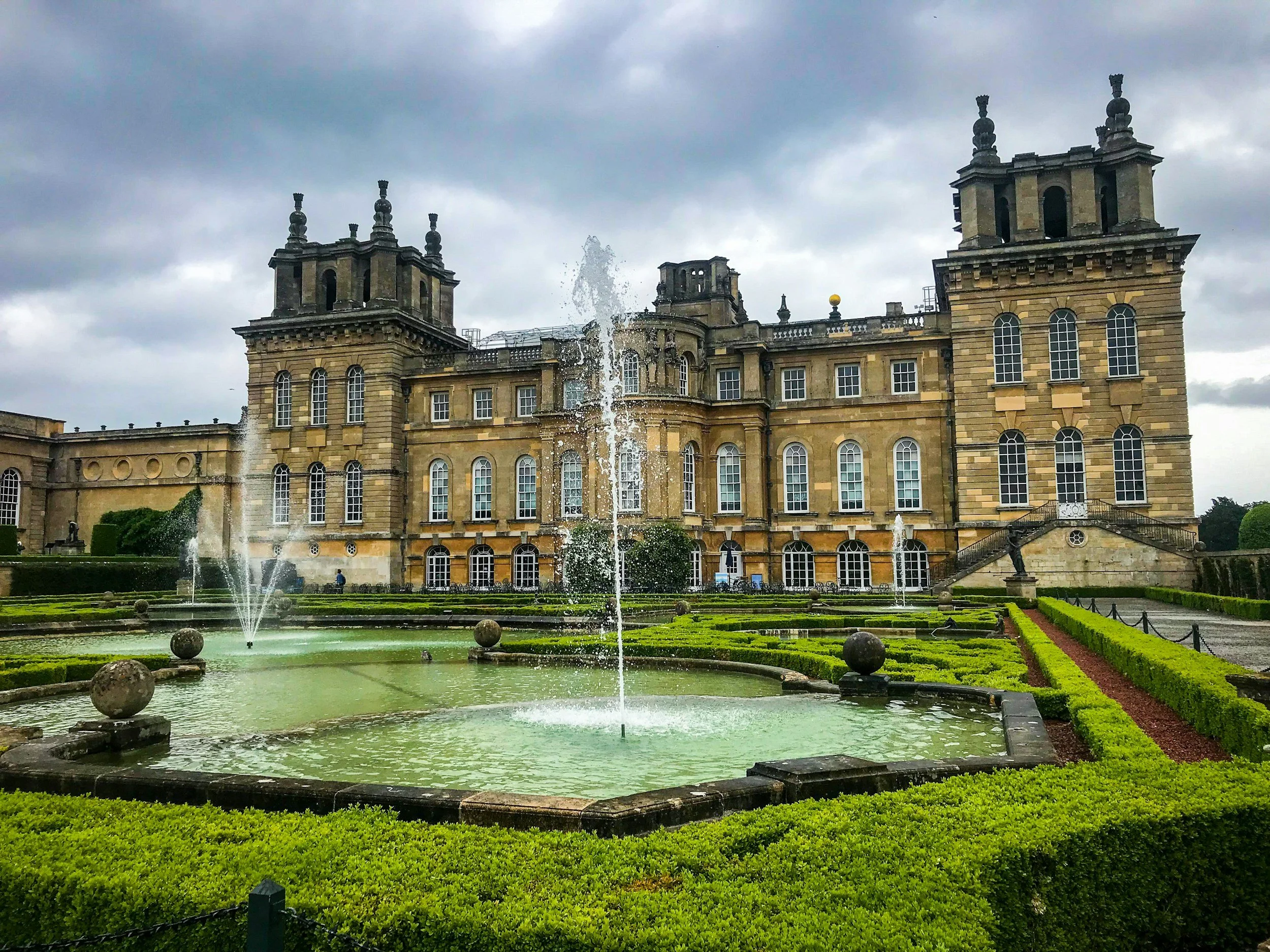 A grand historic castle-like building with multiple windows and towers, surrounded by well-manicured gardens and fountains under a cloudy sky.