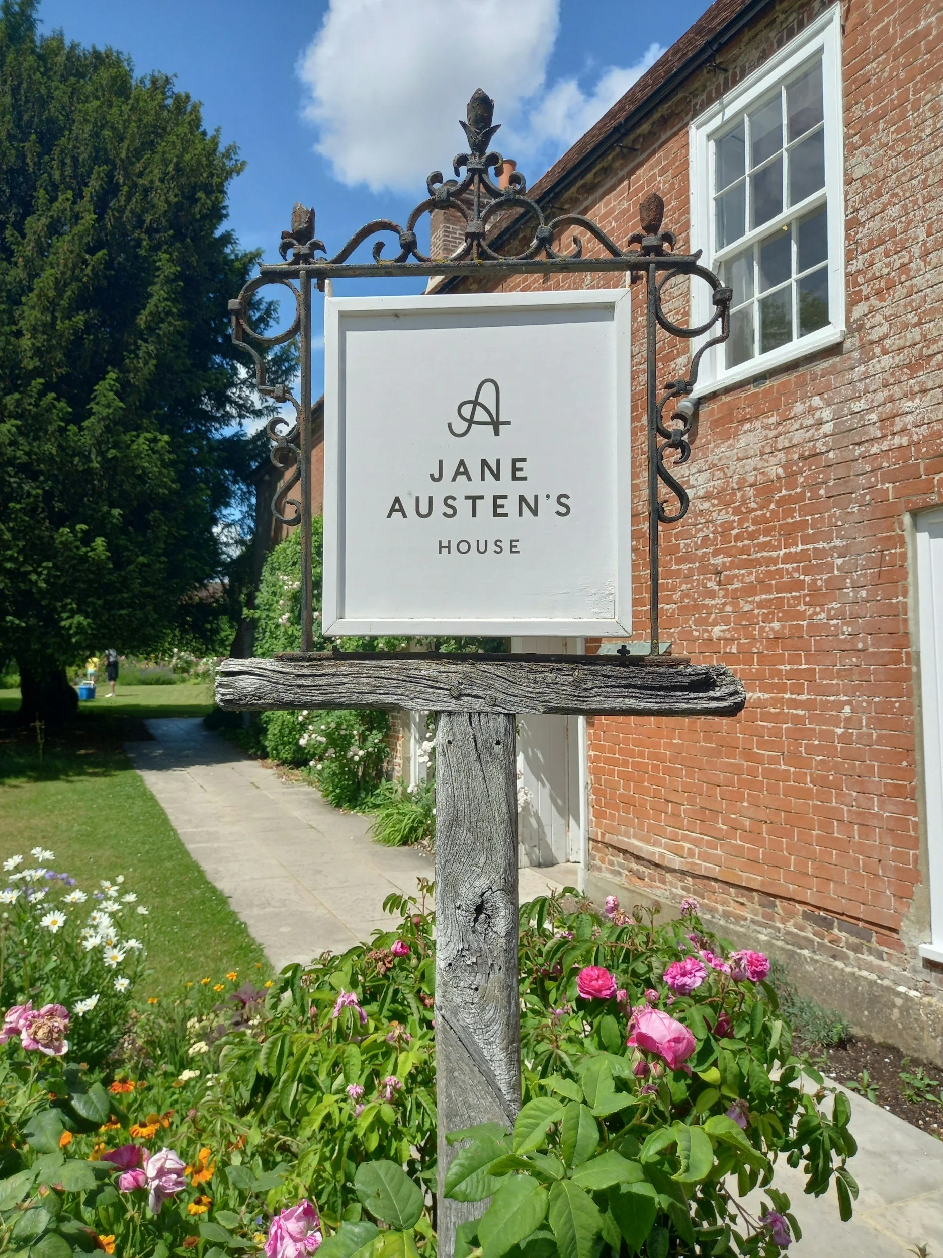 A sign on a wooden post reads 'Jane Austen's House' with a decorative wrought-iron frame, located outside a red-brick house with white trim and multiple windows, surrounded by a garden with colorful flowers and lush green trees under a partly cloudy sky.