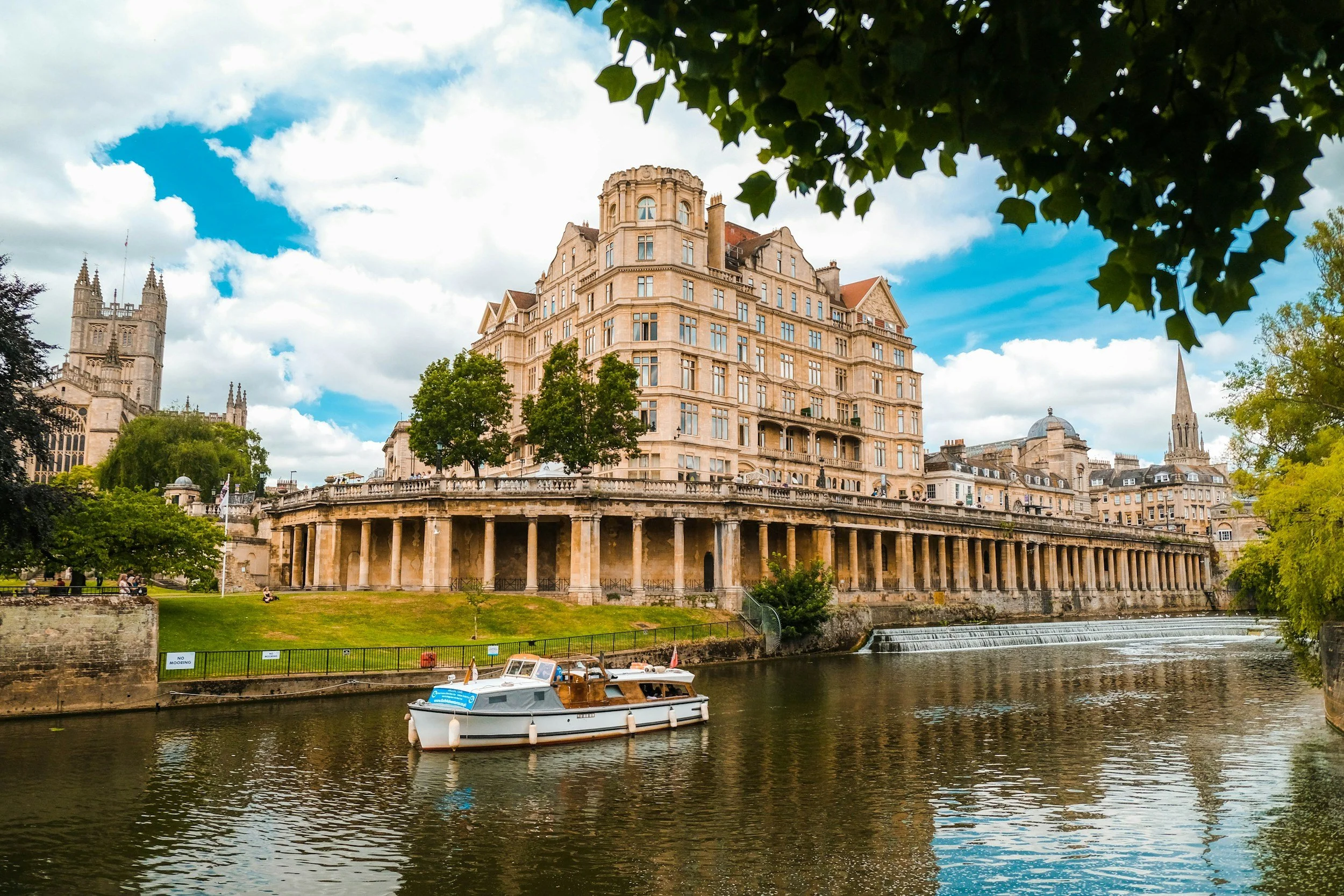 View of a historic building along the river, with trees and a boat in the foreground, and church steeples in the background under a partly cloudy sky.