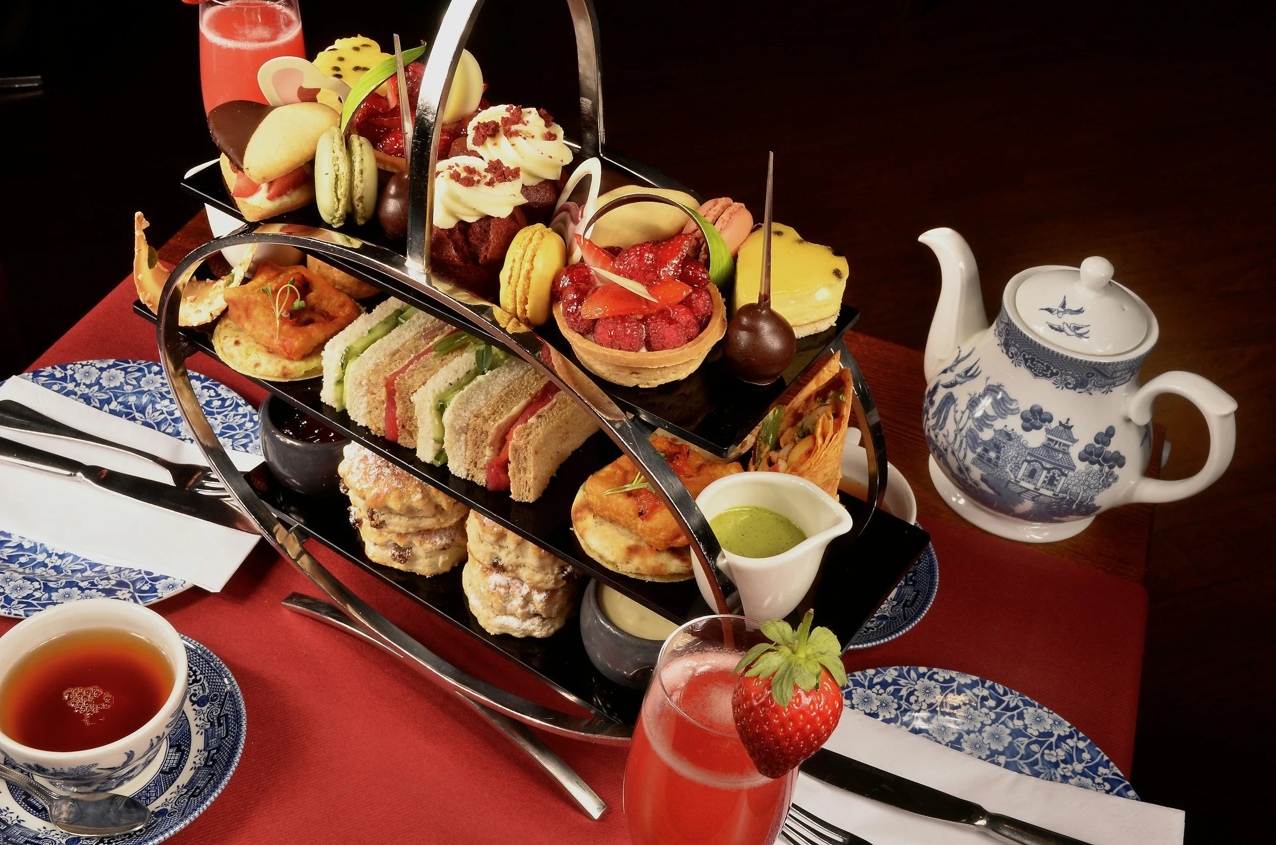 An elegant afternoon tea setup with a tiered tray of assorted desserts, finger sandwiches, scones, and fresh fruit, accompanied by a teapot, teacup, and a strawberry garnish on a pink-colored drink.