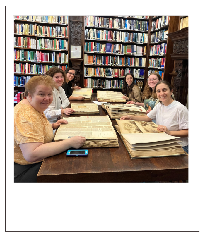 Six people sit around a table in a library, looking at large open books with old pages, with shelves full of books behind them.