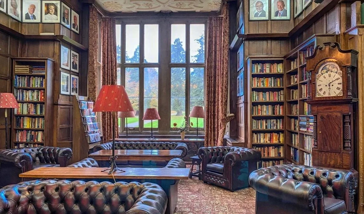 Interior of a classic library with large windows, wooden bookshelves filled with books, leather armchairs, and lamps.