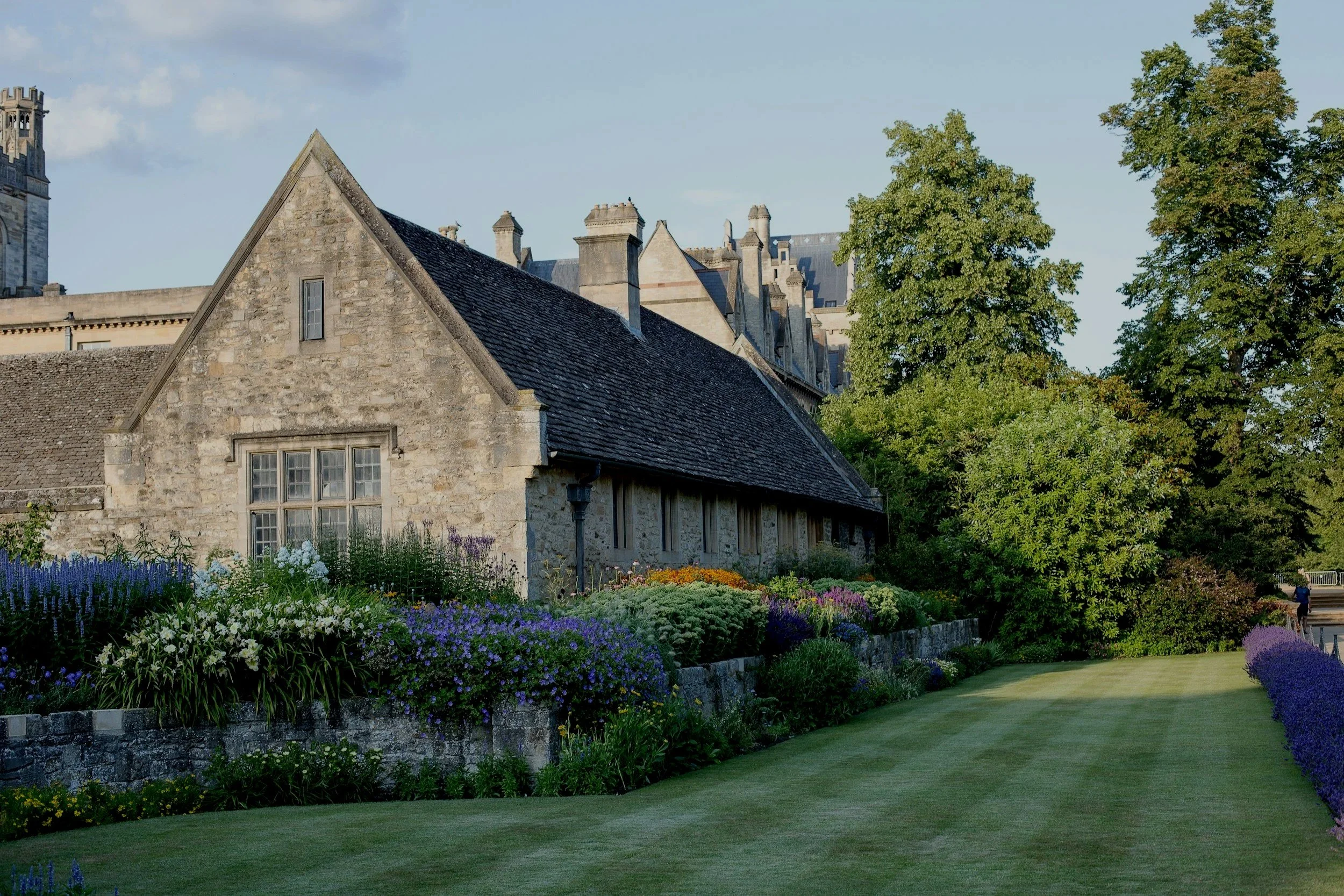 An old stone building with a sloped roof, surrounded by colorful flowers and lush green trees, and a well-maintained grassy lawn in the foreground.