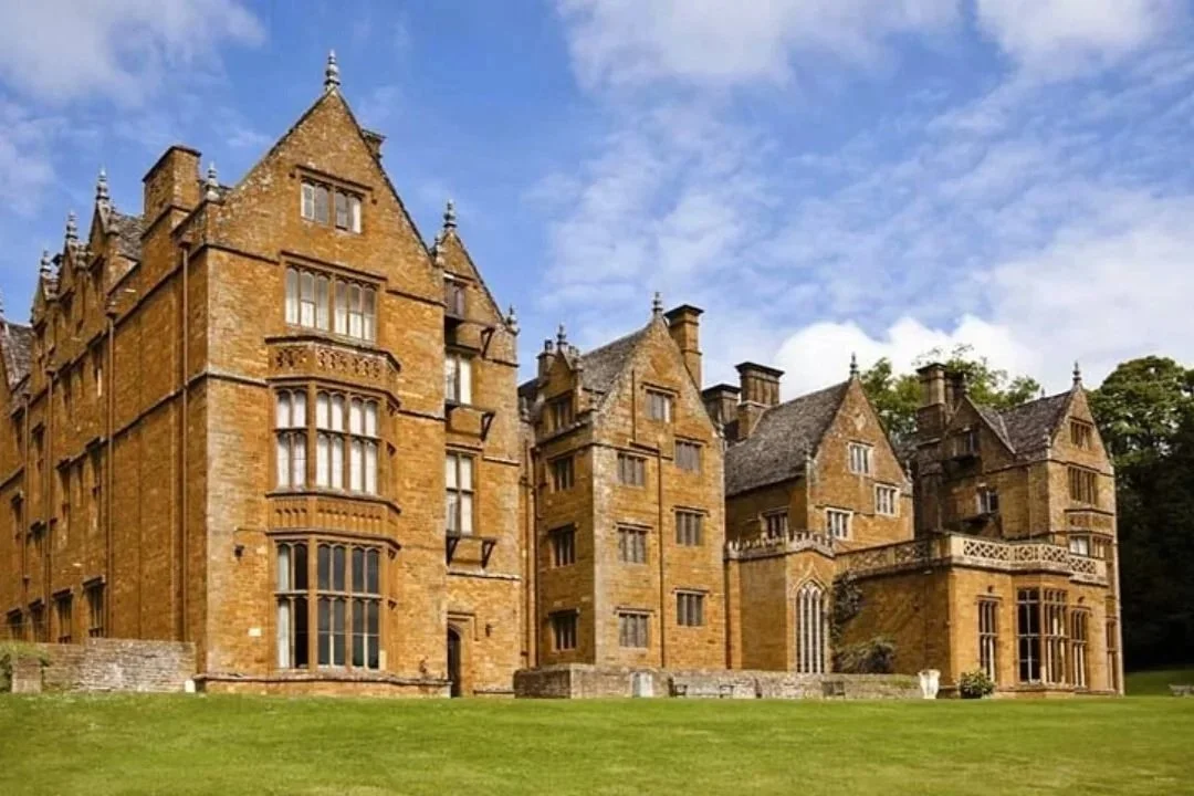 A large, historic castle with stone walls and multiple towers, surrounded by a green lawn and trees under a partly cloudy sky.