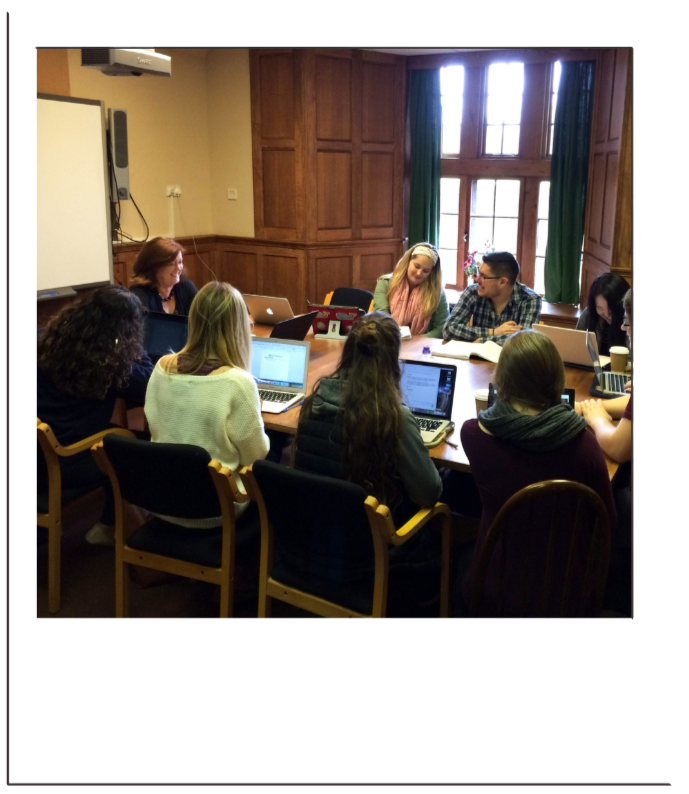 Group of eight young adults sitting around a rectangular conference table in a wood-paneled room with large windows, using laptops and engaged in discussion.