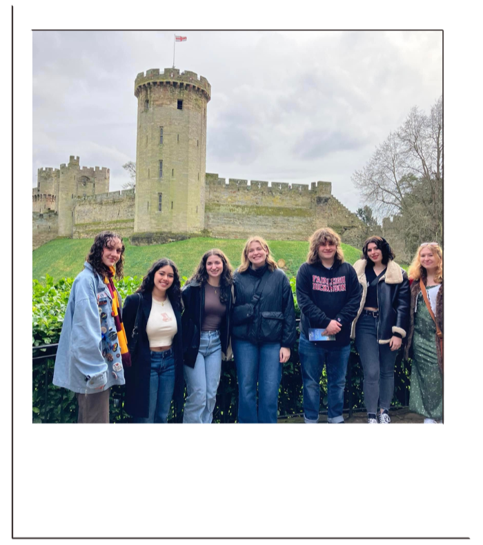 Six young women standing in front of a medieval castle with a flag on top, surrounded by greenery, on a cloudy day.