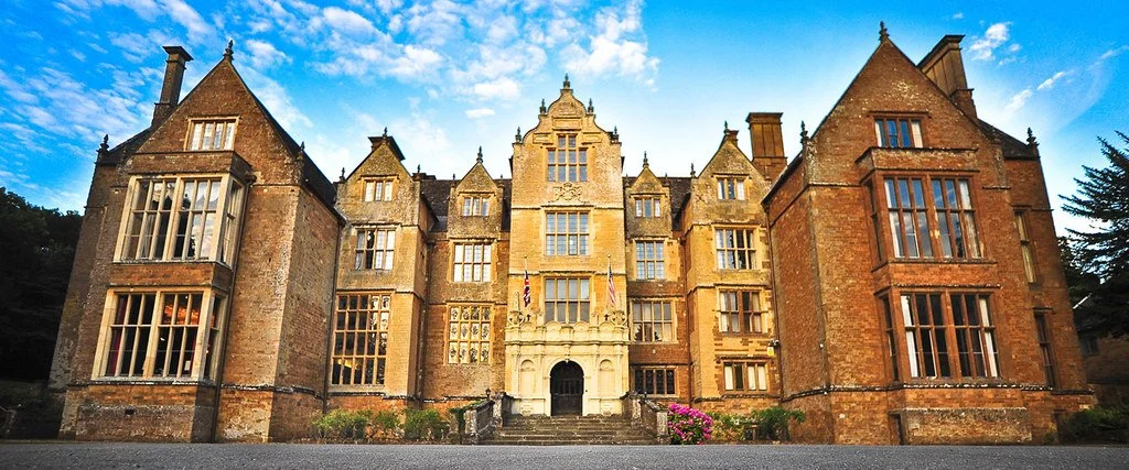 Wroxton Abbey, a large historic abbey with stone walls, multiple towers, and large windows, set against a partly cloudy sky.