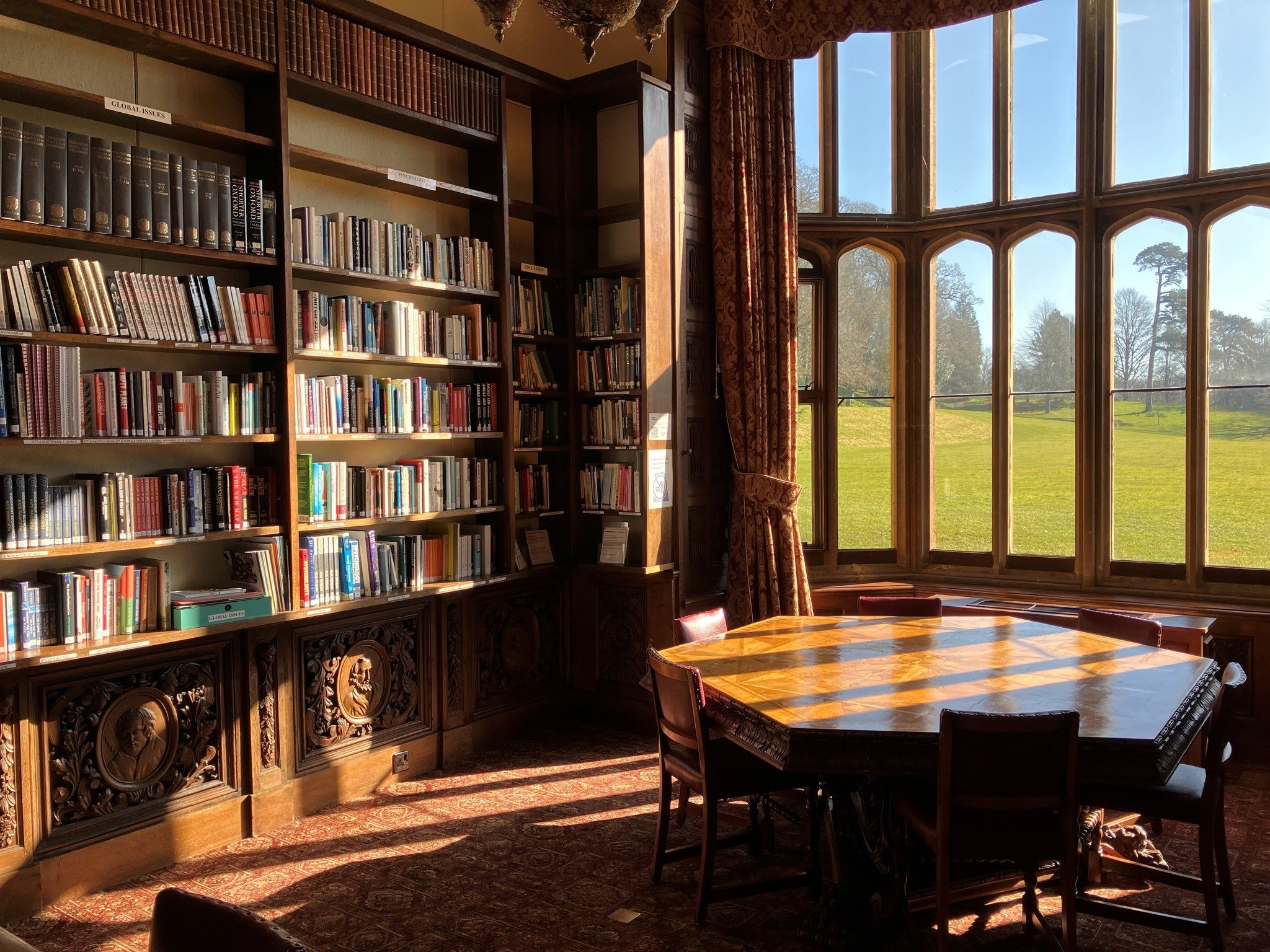 Wroxton College's cozy library room with high wooden bookshelves filled with books, a large wooden table with chairs, and tall windows allowing sunlight to stream in, revealing an outdoor landscape.