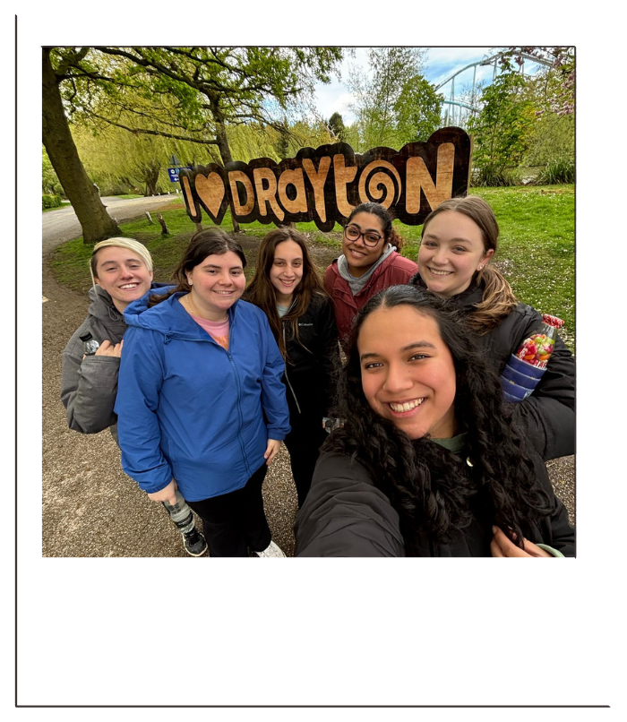 Group of six women standing outdoors in front of a wooden sign that reads 'I ❤️ DAYTON' with trees and a roller coaster in the background.