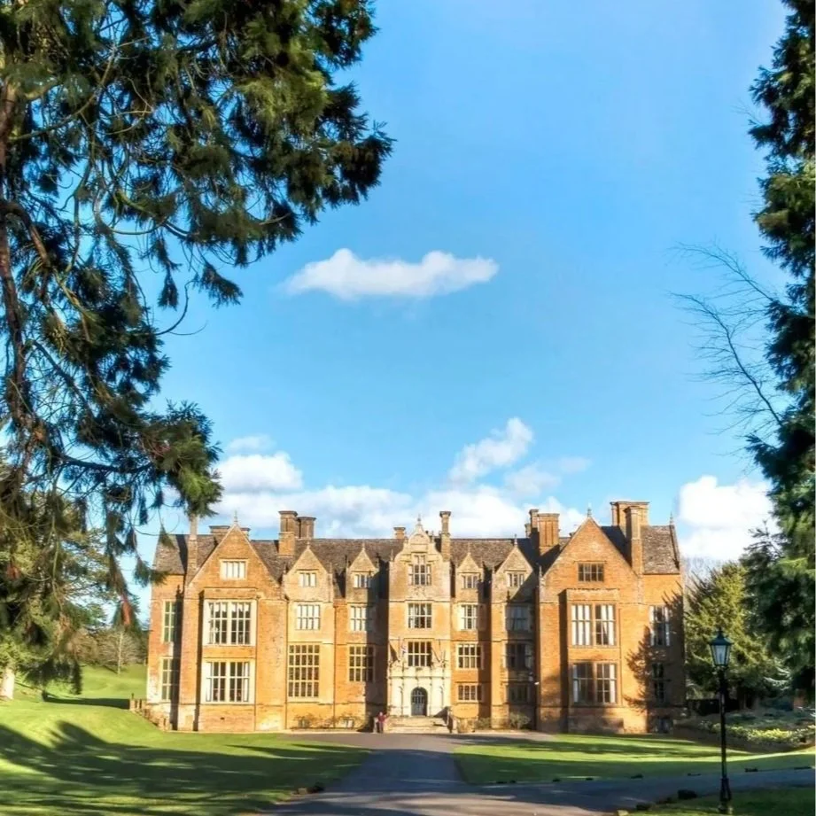 A large, historic brick building with multiple towers and many windows, surrounded by green trees and a well-kept lawn under a partly cloudy blue sky.
