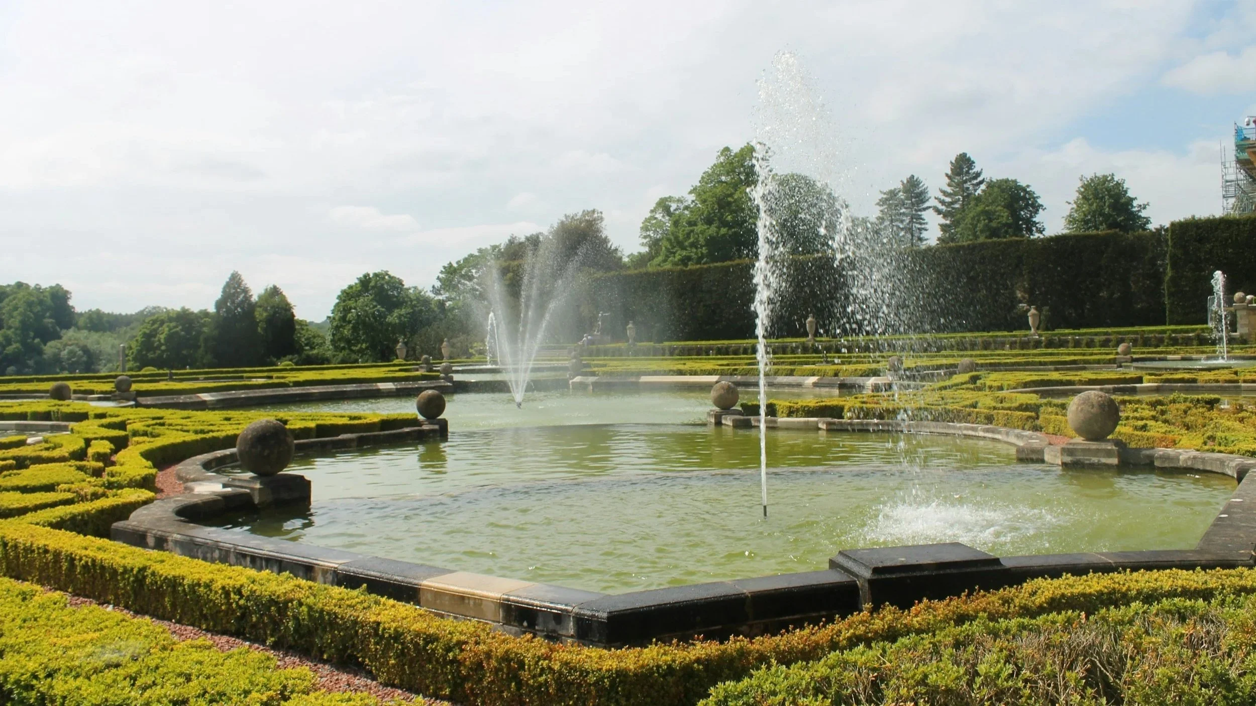 A large outdoor fountain with multiple sprays of water in a landscaped garden with trimmed hedges, trees, and a cloudy sky.