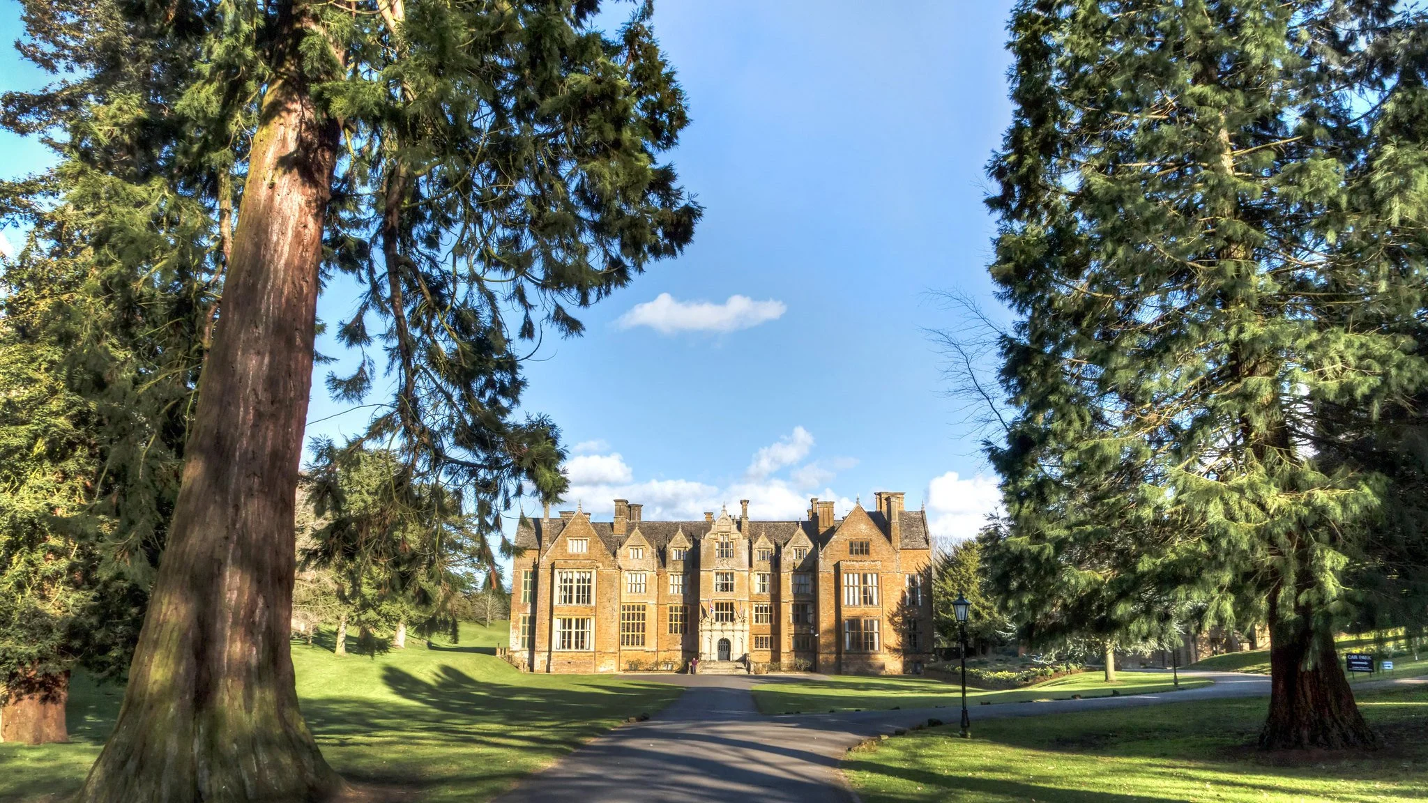 A large historic brick building with multiple chimneys surrounded by greenery and tall trees, under a blue sky with scattered clouds.