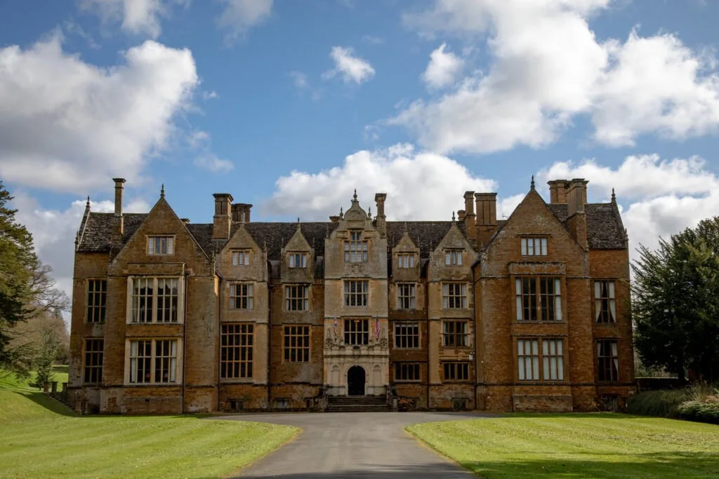 A large historic stone castle with multiple stories and pointed gables, set against a partly cloudy sky and surrounded by green lawns and trees.