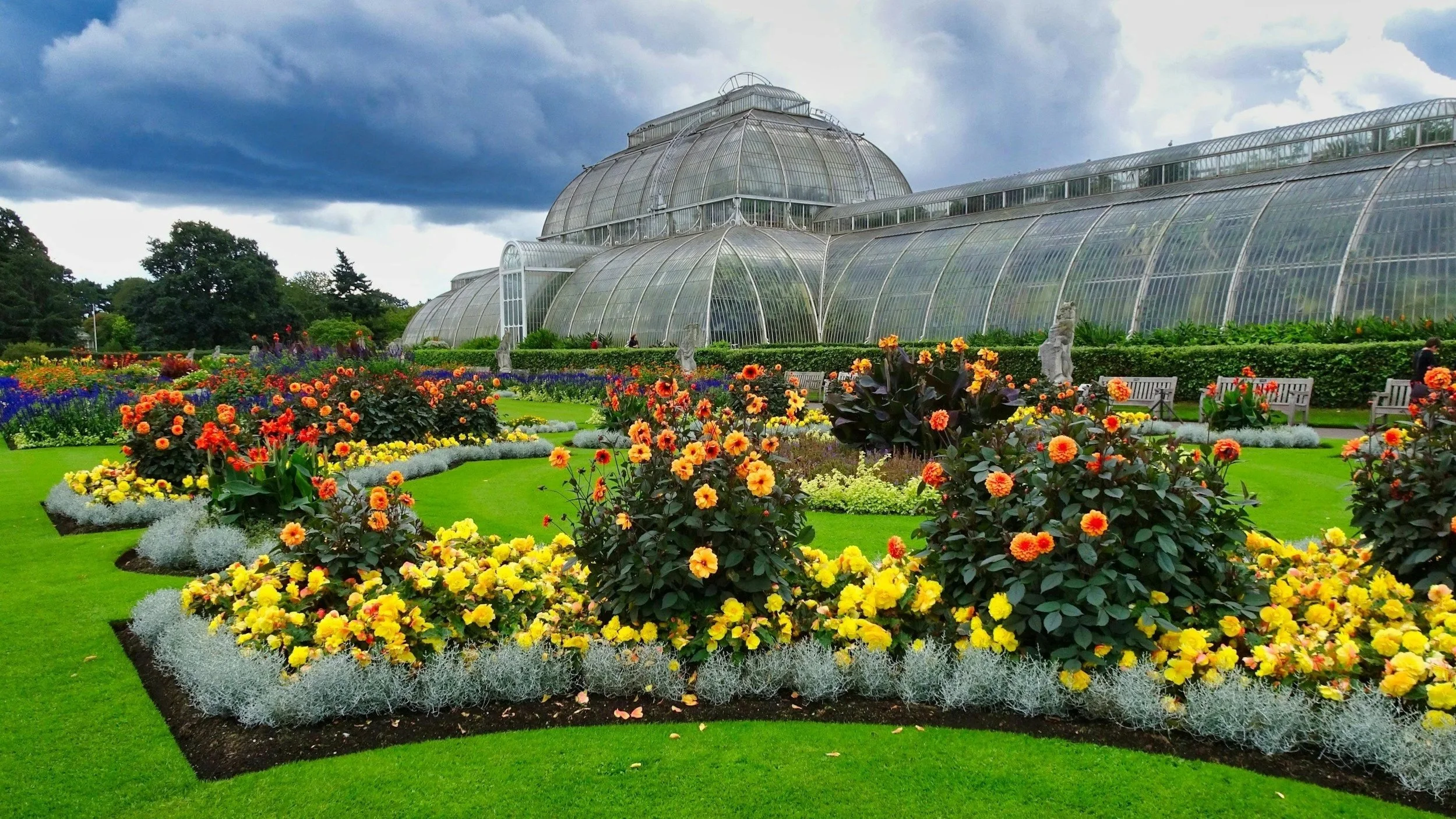A large glass greenhouse surrounded by colorful flower gardens with green grass, statues, and benches under a cloudy sky.