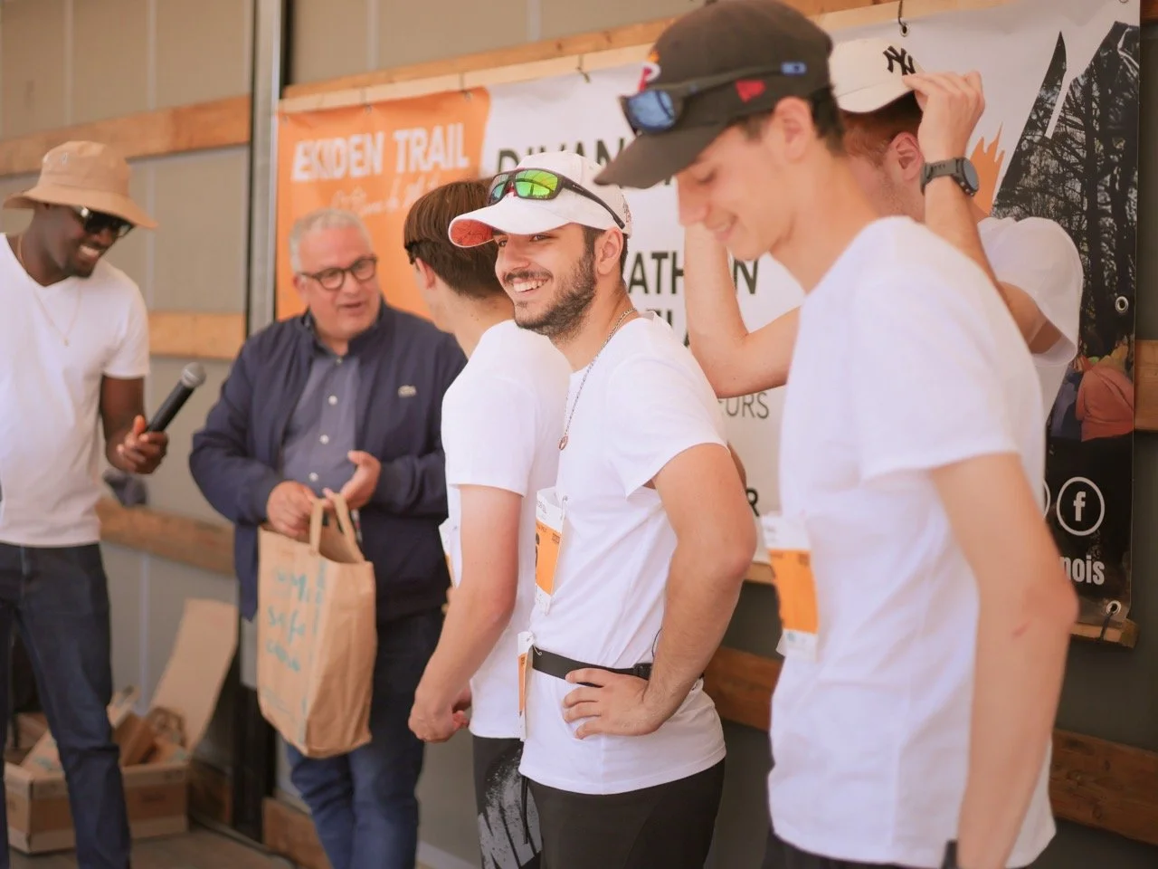 Groupe de jeunes hommes en discussion lors d'un événement, certains portant des casquettes et des lunettes de soleil, avec un homme plus âgé tenant un sac en papier, dans un lieu avec un panneau "Enden Trail" en arrière-plan.
