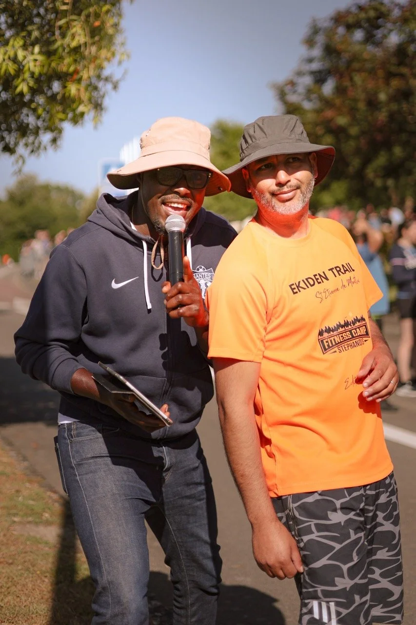 Deux hommes portent des chapeaux et se tiennent côte à côte lors d'un événement en plein air, avec un groupe de personnes en arrière-plan.