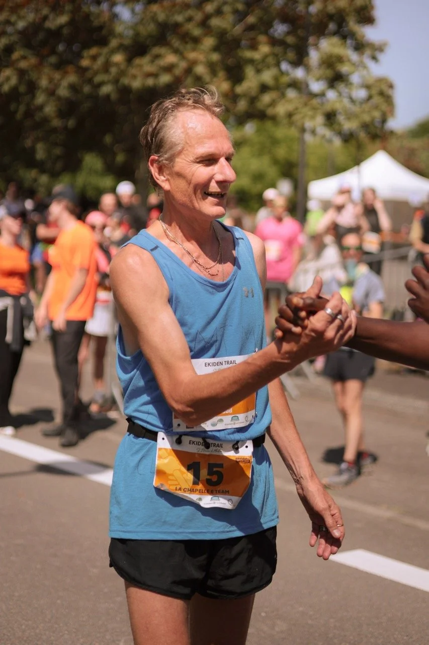 Un homme âgé, souriant, portant un maillot de course bleu et un sport défini, échangeant une poignée de main avec une autre personne lors d'une course ou d'un événement en plein air, avec des spectateurs et des tentes en arrière-plan.
