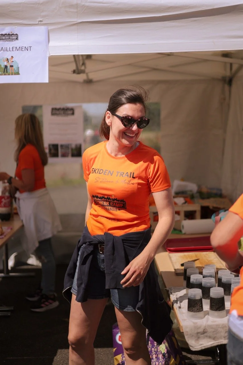 Une femme souriante portant un t-shirt orange, des lunettes de soleil noirs, et un pull attaché à la taille, se tient dans une tente lors d'un événement.