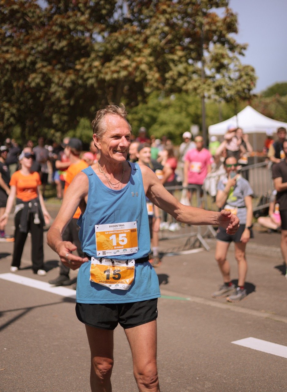 Un homme âgé souriant, portant un maillot bleu et un dossard numéro 15, participant à une course en plein air avec de nombreux autres coureurs devant des arbres et une tente blanche.